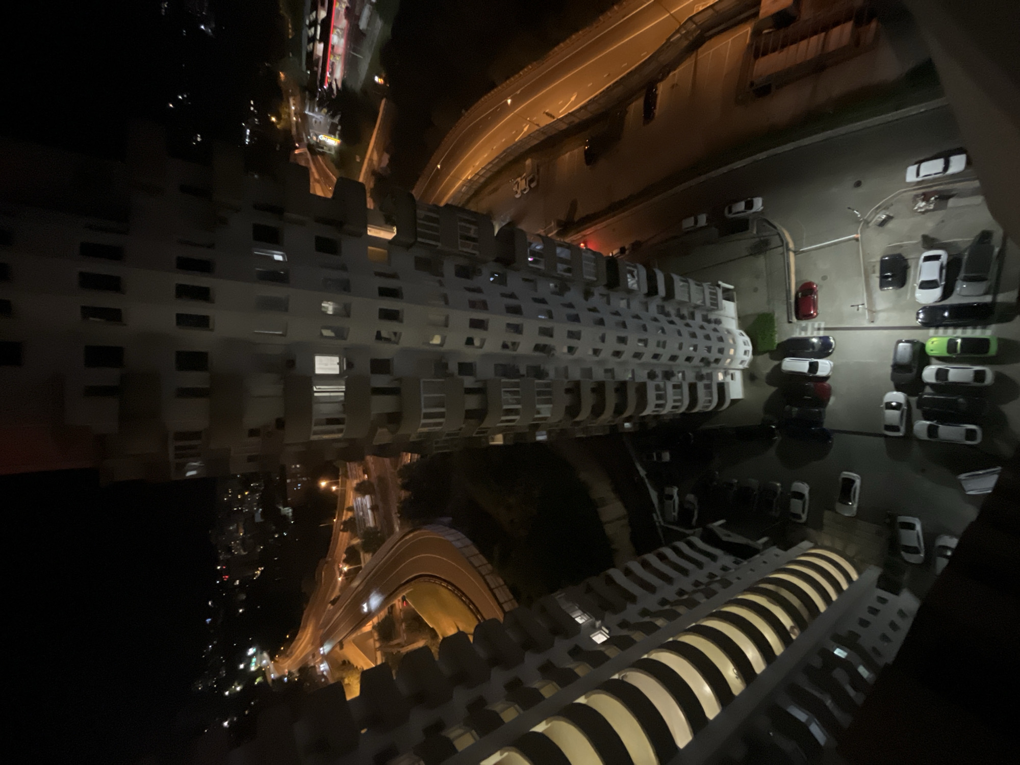 An aerial night shot captures the city of Sochi, Russia, with a focus on high-rise buildings and illuminated roadways. In the foreground, two modern apartment buildings dominate the frame, their balconies and windows casting warm light into the darkness. To the right of the buildings, a multi-lane highway curves through the landscape, its bright headlights and taillights creating streaks of light as vehicles navigate the urban environment. Further into the background, a sprawling cityscape unfolds, dotted with numerous lights indicating residential areas and commercial activity. The overall impression is one of a vibrant, bustling city at night, with the juxtaposition of towering architecture and dynamic transportation infrastructure.