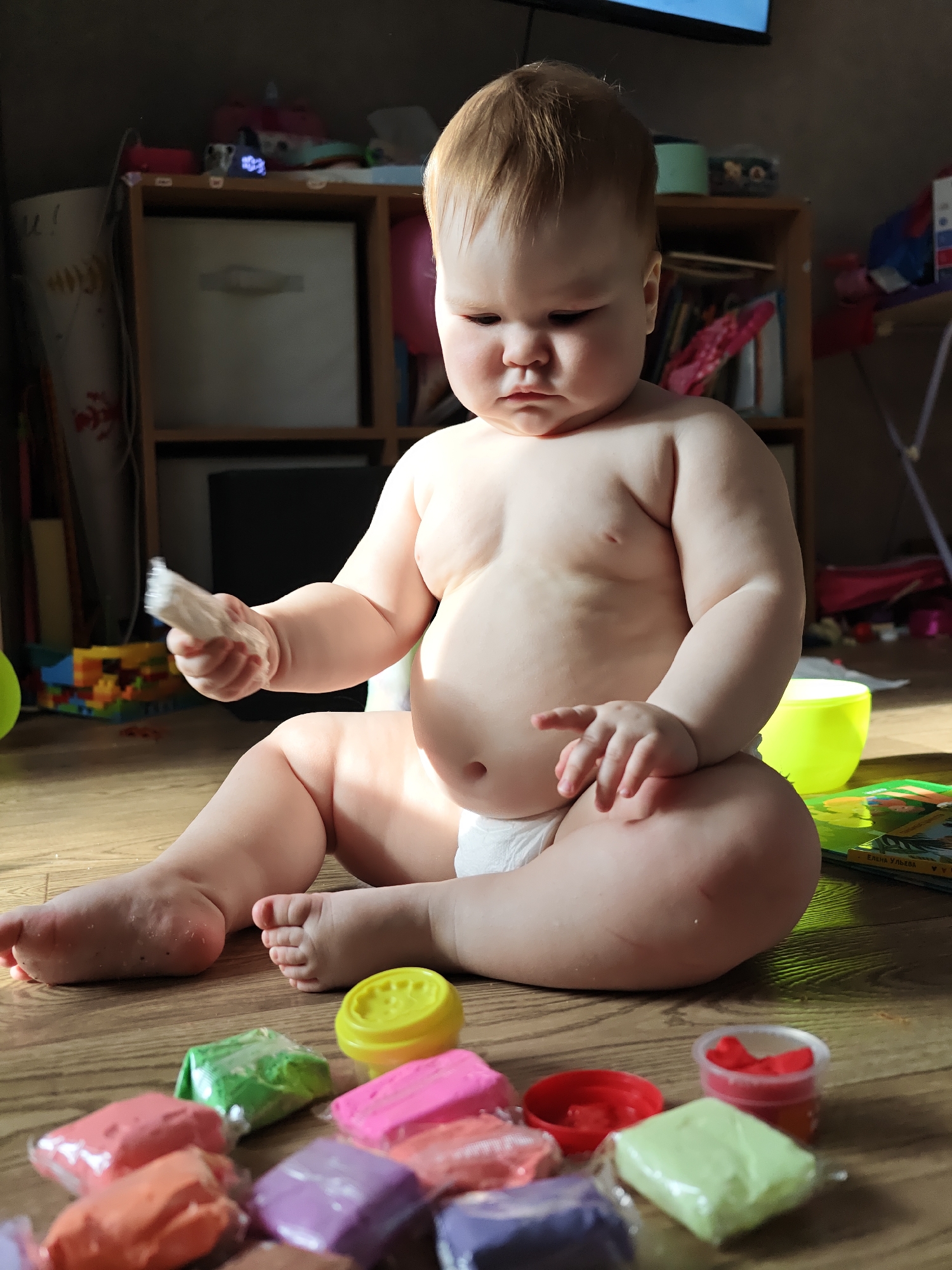 The scene depicts an infant seated on a wooden floor in an indoor residential setting. The specific location within Chaikovsky, Russia, cannot be determined from visual cues in the image. The infant, appearing to be a toddler with light brown hair, is wearing a white diaper and is positioned centrally, facing slightly left with a neutral to pensive expression. The infant's left hand holds a small, elongated, light-colored object, possibly a piece of white modeling clay, while the right hand rests on the right thigh.
Numerous individually wrapped blocks of modeling clay in various colors (orange, pink, purple, green, light green, brown) are scattered across the wooden floor in the foreground and around the infant. Several small open plastic containers, including yellow and red lids, and one translucent container holding red clay, are also present. A green plastic cup and a children's book, partially displaying the author's name "Елена Ульева," are situated to the infant's right.
In the background, a light brown wooden shelving unit contains various items, including a light-colored fabric storage cube in one compartment. A small digital clock displaying "10:23" is visible on the top shelf, alongside other miscellaneous objects and books. A dark rectangular object, possibly a television screen, is mounted above the shelving unit. Colorful building blocks are visible on the floor in the left background. The room is illuminated by natural light originating from the left, creating highlights and shadows on the subjects and environment.