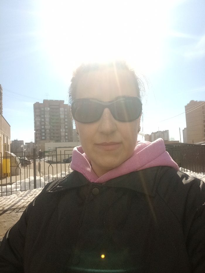 A female individual is depicted in a close-up outdoor portrait, likely a selfie, in Serpukhov, Russia, on a bright, sunny day. The subject is positioned centrally, facing directly towards the camera, and exhibits a neutral expression with closed lips. She wears dark-tinted, wrap-around sunglasses, a black collared jacket with visible snap buttons on the front, and a pink hooded sweatshirt underneath. Her hair, appearing dark, is pulled back from her face.
The background reveals an urban environment under a clear blue sky. A prominent lens flare from the sun is visible in the upper right quadrant of the image. In the mid-ground, patches of melting snow are present on the ground. A dark metal fence, possibly for pedestrian separation or security, extends horizontally across the scene, behind which indistinct parked vehicles are visible on what appears to be a street or parking area. To the far left, a bright yellow utility box or container is partially visible.
Multiple multi-story apartment buildings, characteristic of urban architecture, are discernible in the background. A large, brick-colored building with numerous windows stands directly behind the subject's left side, while another lighter-colored, taller building is partially visible further to the right. The scene is brightly lit by direct sunlight, casting shadows and creating high contrast.
