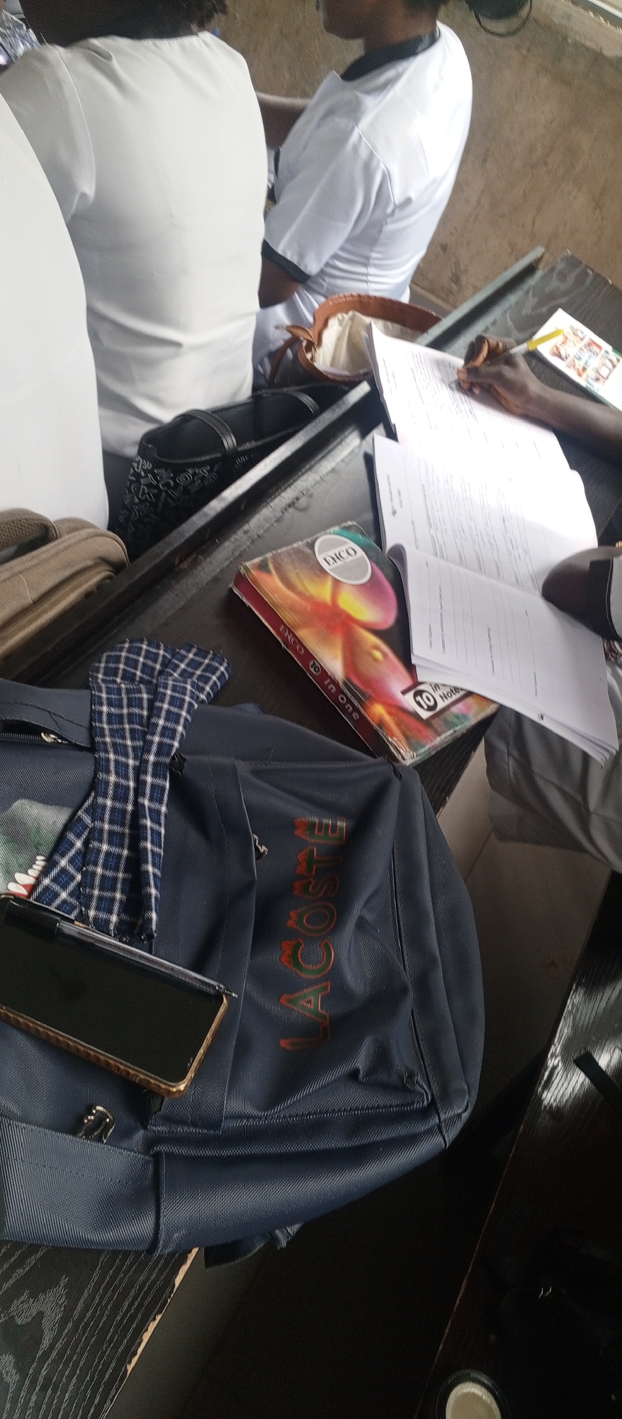 This image depicts a classroom setting, likely in Kuson, Nigeria, with a focus on educational materials and student belongings. In the foreground, a dark blue backpack with the "LACOSTE" logo is prominent. A blue and white checkered cloth is placed on top of the backpack. A smartphone lies on the backpack's surface. To the right of the backpack is a book titled "ENCO 10 In One". In the background, two individuals, likely students, are seated at a table. One is wearing a white top, and the other is also in white attire, with their hands engaged with papers. The environment appears to be a study area, possibly a classroom or a communal study space, with a wooden table surface and a neutral-colored wall in the background. The overall scene suggests a moment of academic activity.