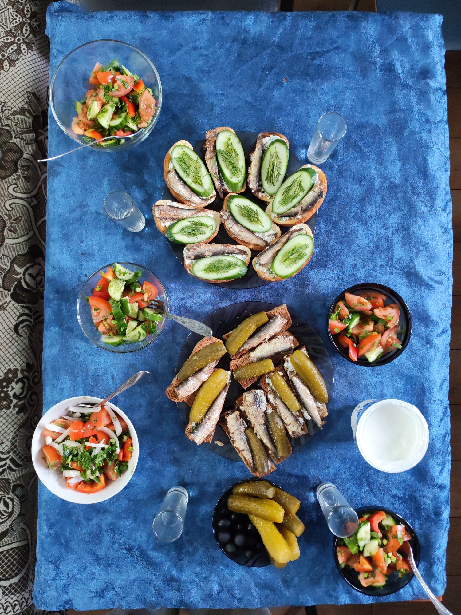 An overhead shot captures a table set with a variety of traditional Russian and Eastern European dishes, suggesting a casual meal or gathering in Shcheleiki, Russia. The table is covered with a textured, deep blue tablecloth.
Central to the spread are two plates of open-faced sandwiches, commonly known as "buterbrody." The top plate features sandwiches made with light-colored bread, topped with what appear to be sprats or sardines, a creamy spread (possibly butter or cream cheese), sliced fresh cucumbers, and sprigs of fresh dill. The bottom plate displays similar sandwiches on dark rye bread, garnished with whole pickled cucumbers and also fish.
Accompanying the sandwiches are several bowls of fresh vegetable salads. There are at least three bowls containing diced tomatoes and cucumbers, some with chopped green onions or herbs. One white bowl also contains sliced white onions mixed with tomatoes and herbs. Three clear, empty shot glasses are strategically placed around the table, hinting at the consumption of spirits, likely vodka, a common pairing with such snacks. A white plastic container, possibly holding sour cream or yogurt, is visible near the bottom right. A small black bowl filled with whole pickled cucumbers is also present.
The lighting appears bright and natural, suggesting it is daytime. On the left side of the frame, a portion of a patterned, possibly upholstered, surface is visible, indicating the table might be placed next to a couch or a similar piece of furniture in an indoor setting. There is no visible text in the image.