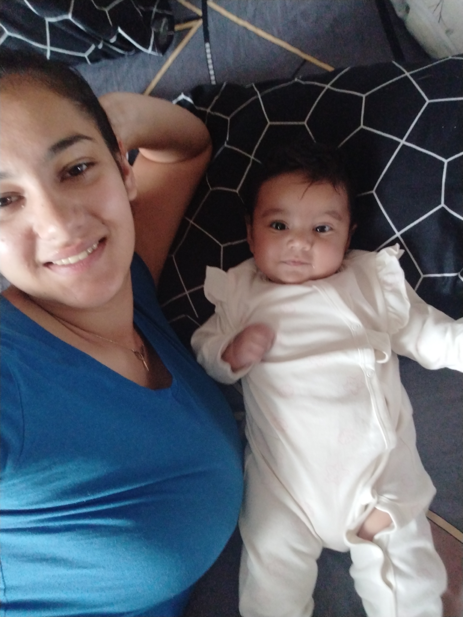 A woman and a baby are lying on a bed in an indoor setting. The woman, wearing a blue t-shirt and a gold necklace, is smiling towards the camera while resting her head on her hand. Next to her, a baby wearing a white onesie is lying on a patterned pillow. The location is Unknown, Unknown.