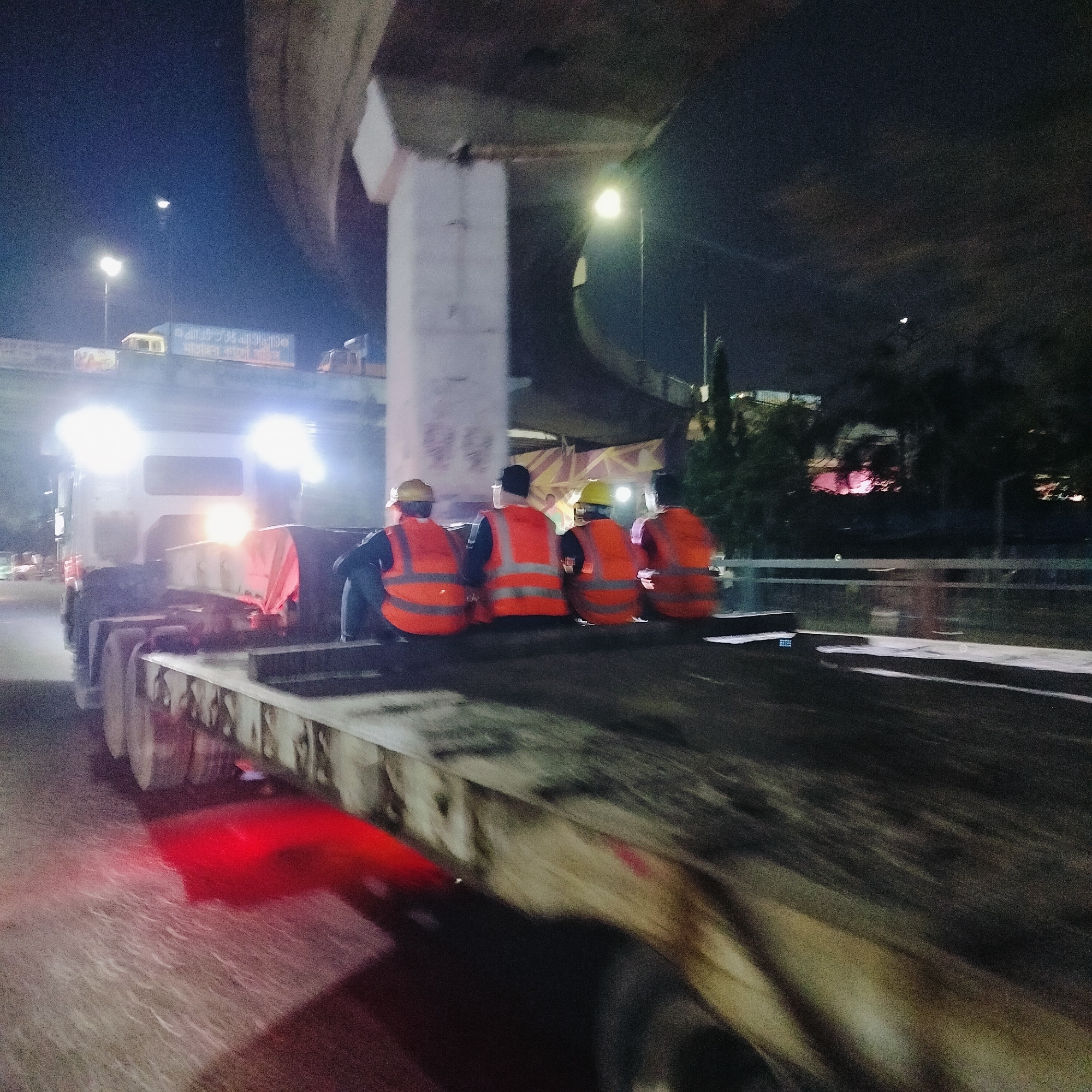 At night, under a bridge support in Dhaka, Bangladesh, four workers in orange vests and hard hats sit on a flatbed trailer attached to a truck. The truck's headlights are illuminated, and red lights shine beneath the trailer. Streetlights are visible in the background. The scene appears to be related to construction or road work.