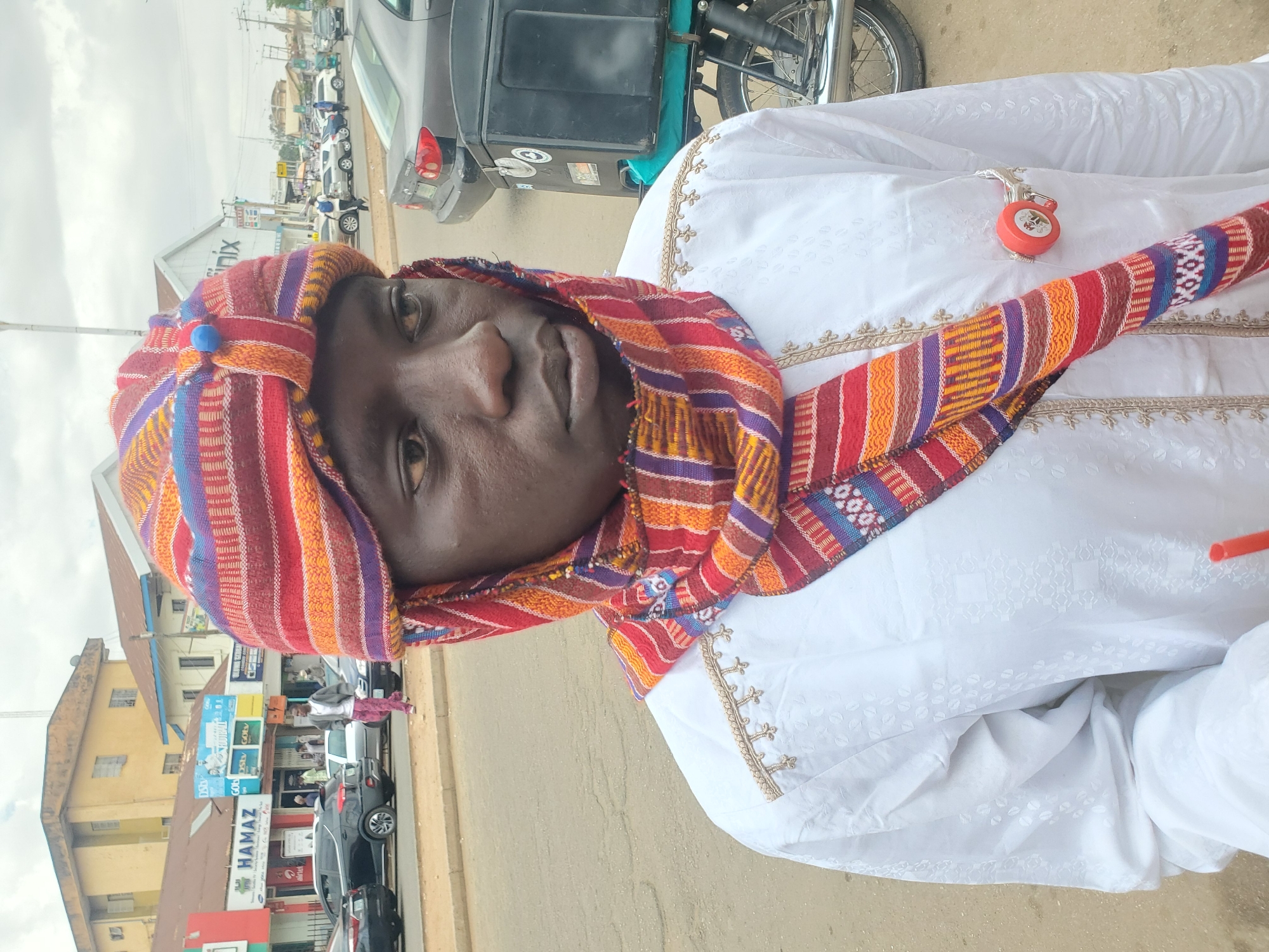 This vertically oriented image, rotated 90 degrees clockwise, captures a person dressed in traditional attire on a street in what appears to be an urban area of Jos, Nigeria.
The main subject is a person, likely male, with dark skin and a neutral expression, looking directly at the camera. They are wearing a pristine white, long-sleeved garment, possibly a caftan or similar tunic, featuring intricate gold-colored embroidery along the neckline and cuffs. A vibrant, multi-colored striped head wrap or turban, primarily in shades of red, orange, yellow, blue, and purple, adorns their head and drapes around their neck like a scarf. A small blue button-like embellishment is visible on the headwrap near the forehead. Clipped to the white garment on the chest area is a small, red, circular object with a silver clasp, possibly a keyring or a novelty item.
The background depicts a bustling street scene with several buildings, some appearing to be storefronts. One building has a reddish-brown corrugated metal roof. Various vehicles, including cars, a motorcycle, and what looks like a tricycle (keke napep), are visible on the street. The sky is overcast, suggesting daytime with diffused lighting.
Visible text on signs in the background includes "NIX" (partially visible, likely part of "Phoenix"), "HAMAZ", "DStv", "GOtv", "C O M", and "Airtel", indicating commercial establishments. The overall impression is that of a candid shot in a lively, commercial district.
