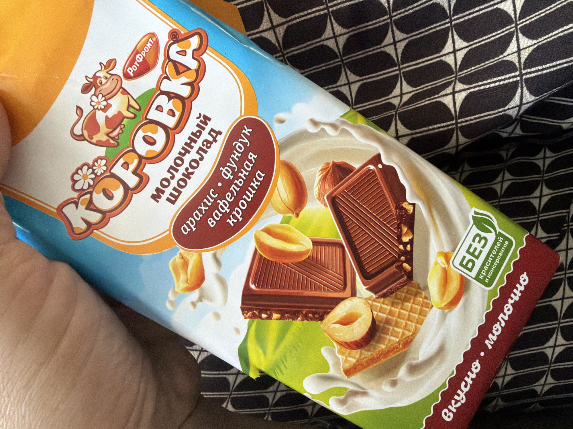 The image shows a close-up of a chocolate bar wrapper with a hand holding it. The wrapper is blue, orange, and green, featuring a cartoon cow with flowers, and images of chocolate, wafers, nuts, and milk splashes. Russian text on the wrapper reads "Коровка" (Korovka), "МОЛОЧНЫЙ ШОКОЛАД" (Milk Chocolate), and other descriptive words. A green and white label indicates "БЕЗ красителей и консервантов" (Without dyes and preservatives). The background is a patterned fabric. No specific location in Mezhevoy, Russia, can be determined from the image.