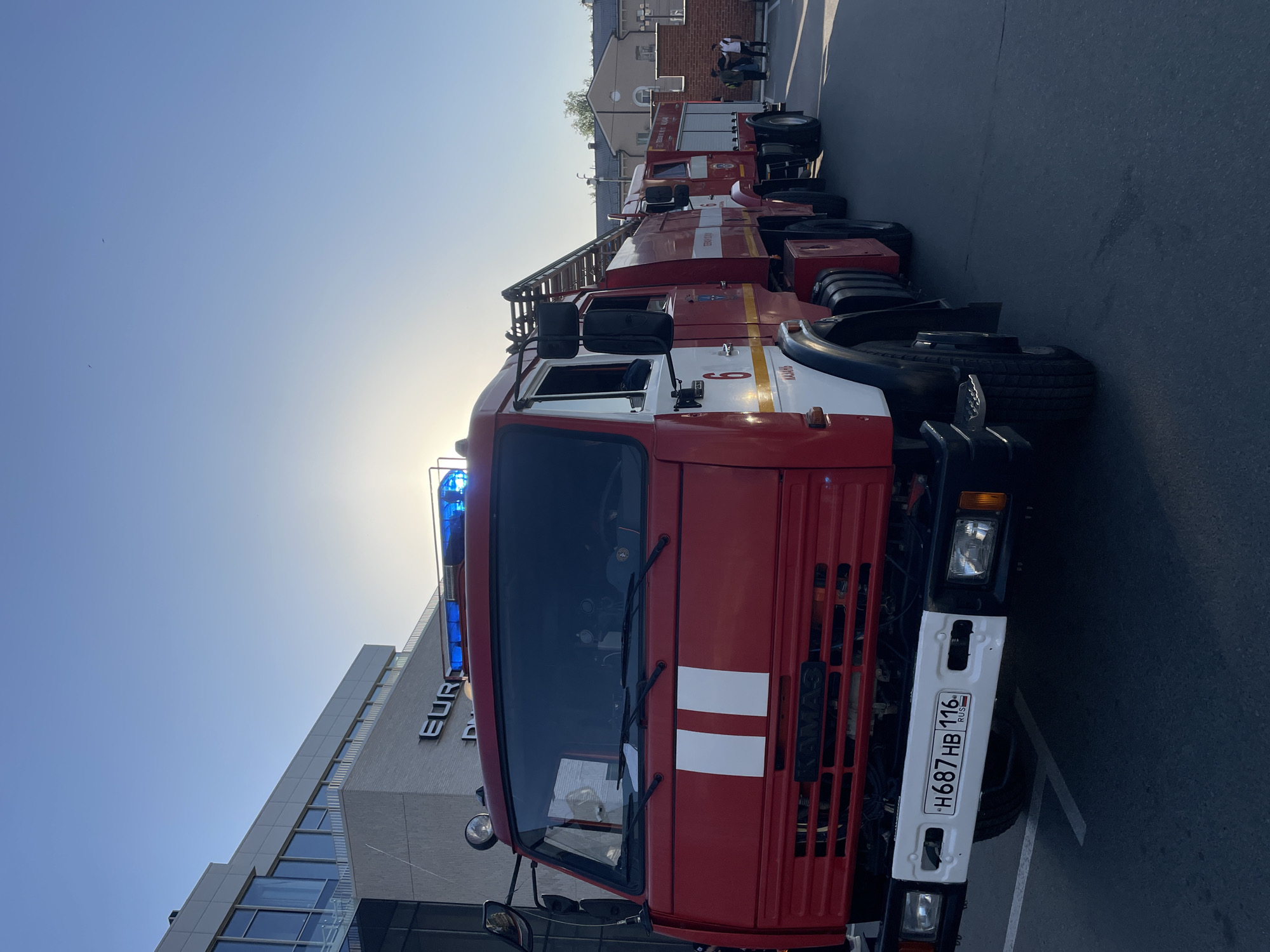The image shows a line of red and white fire trucks parked on an asphalt surface in Kazan, Russia. The front of one of the trucks is prominent, featuring a blue emergency light on the roof, a white grill with two vertical white stripes, and a Russian license plate with the code "H687HB 116". A modern building with glass windows and the word "EURO" visible on its façade stands behind the trucks. Further in the background, a brick building and several people are visible. The sky is clear and bright, suggesting a sunny day.