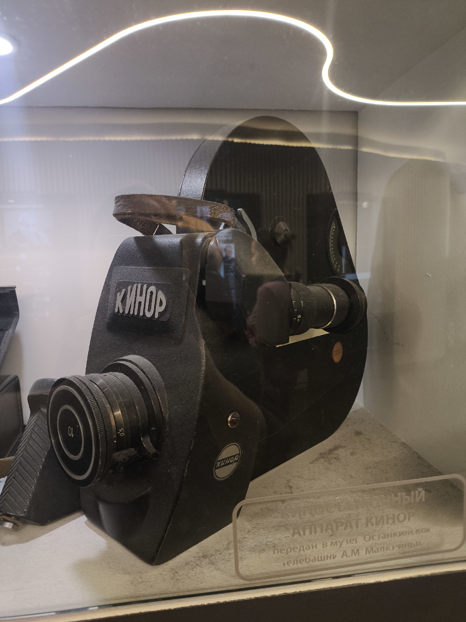 The image shows a vintage "Kinor" brand film camera displayed in a glass case. A descriptive plaque is placed near the camera, stating "Cinematographic apparatus 'Kinor' given to the Ostankino TV tower A.M. Manarin". The camera is black with various dials and lenses visible. The background appears to be a museum exhibit, likely located in Moscow, Russia, given the reference to the Ostankino TV tower, which is situated in Moscow.