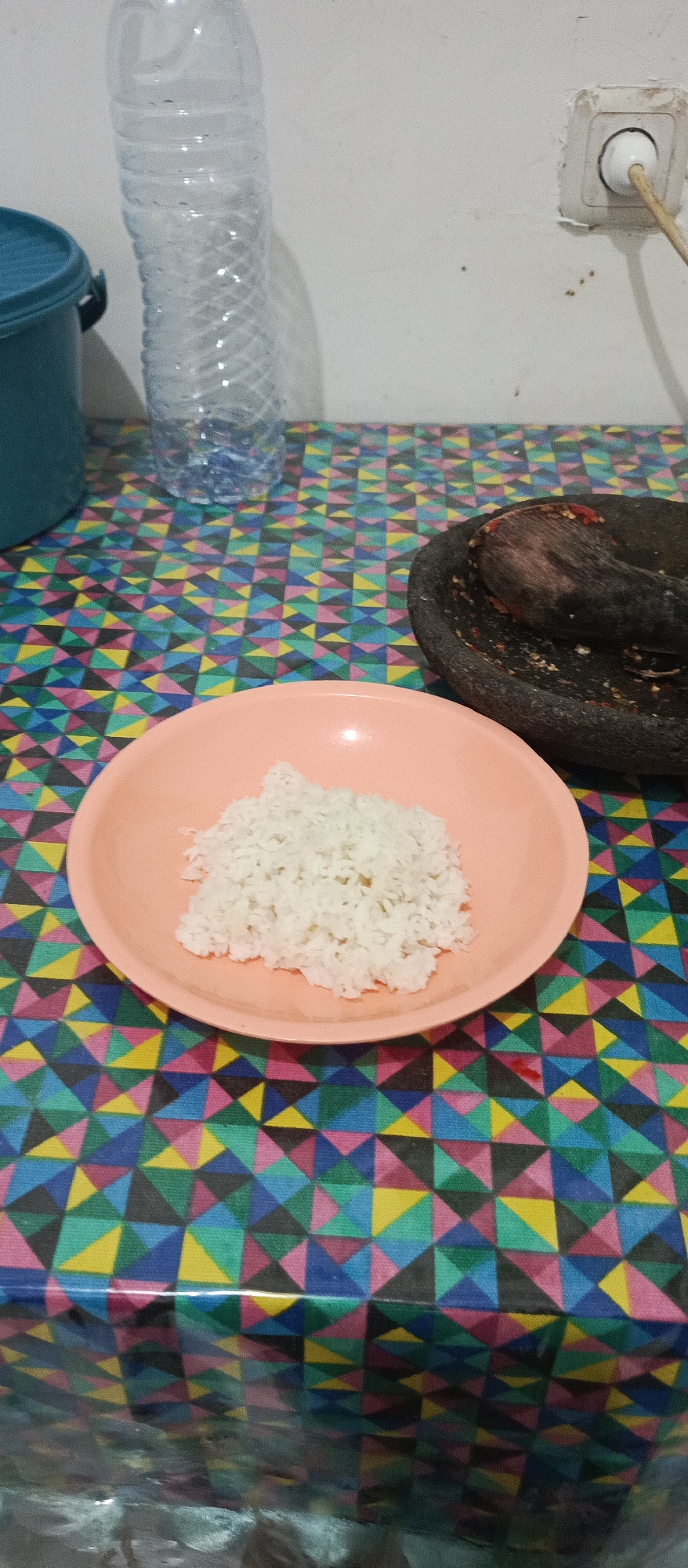 The image depicts a close-up, overhead view of a table covered with a colorful, geometric-patterned tablecloth. On the table, a pink bowl contains a pile of white rice. To the right of the bowl is a dark-colored mortar and pestle, which appears to have some red chili residue inside. In the background, a clear plastic water bottle stands upright. To the left of the bottle, a portion of a teal-colored bucket or container is visible. The background wall is white, with an electrical outlet and a visible cord. The scene appears to be a domestic setting, possibly a kitchen or dining area. The image does not contain information that can be used to determine the specific location within Arjasa, Indonesia.