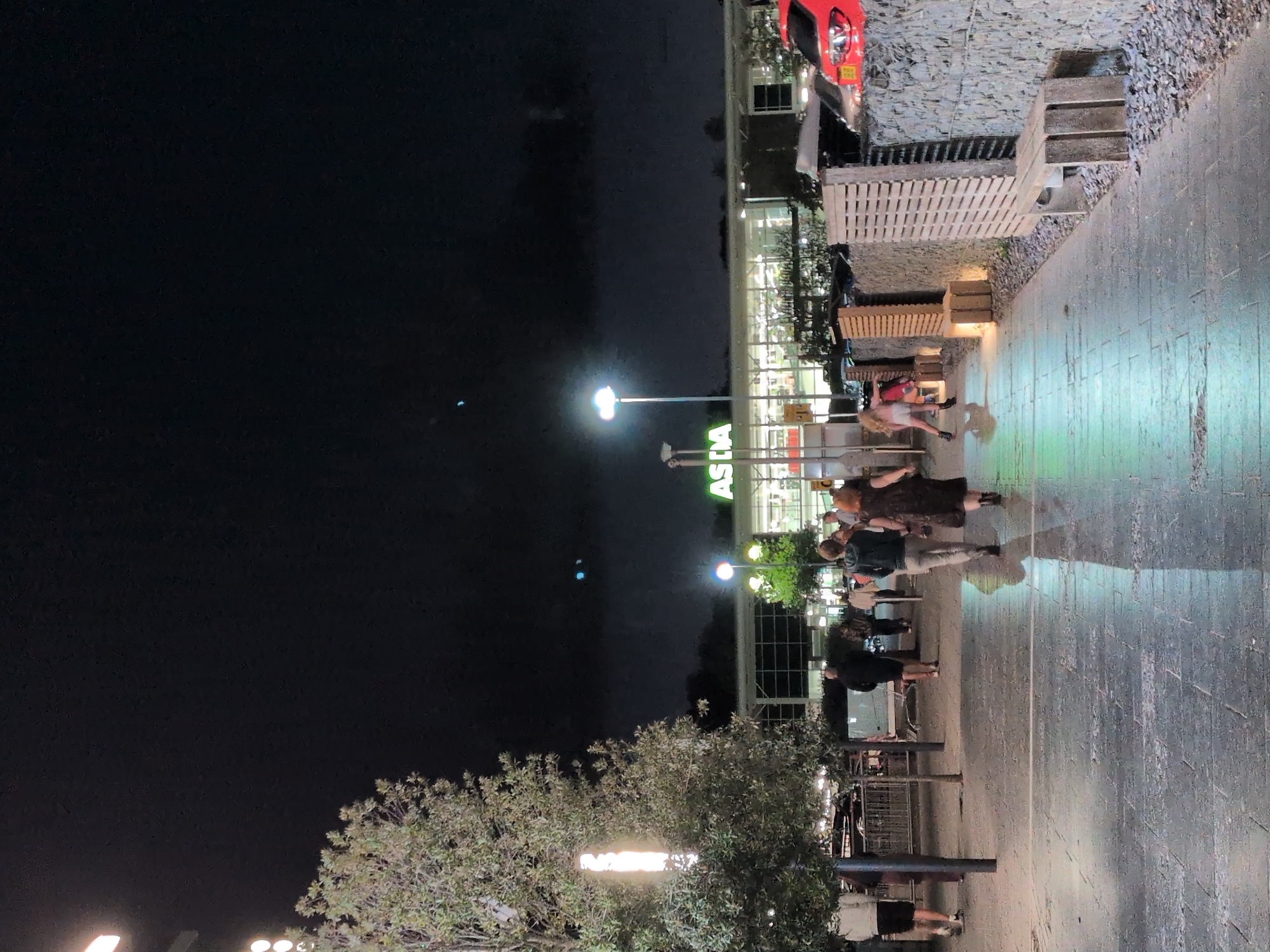 The image shows an outdoor scene at night, likely in a public area with a building featuring illuminated signage for "ASDA". Several individuals are walking on a paved pathway. A large tree with foliage is visible on the left side of the frame. Streetlights illuminate the area, casting reflections on the wet pavement. The background shows the ASDA building and parked vehicles, including a red car. The scene appears to be a shopping area or retail park.