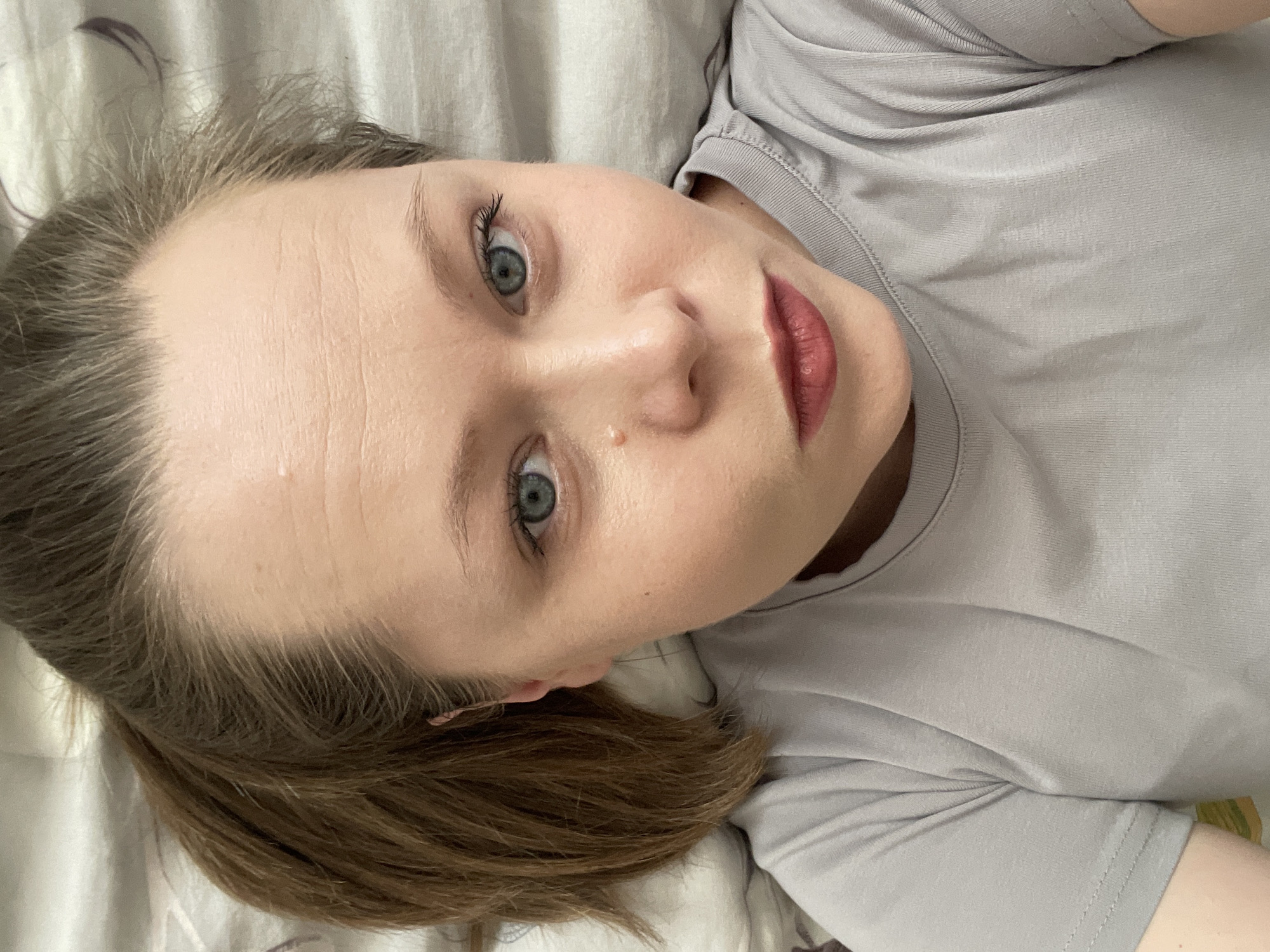 This is a close-up, slightly overhead portrait of a young woman lying on her back, looking directly at the camera. Her head is tilted to her left, with her light brown hair spread around her.
The subject has fair skin, prominent blue-green eyes, and light brown hair. Her forehead exhibits some fine horizontal lines. There is a small, light-colored bump or mole visible on her right cheek, below her eye. She is wearing light makeup, including mascara that enhances her eyelashes and a reddish-pink lipstick on her full lips. Her expression is neutral and calm, with a direct gaze.
She is wearing a plain, light gray t-shirt with a rounded neckline. The fabric of the t-shirt shows some minor wrinkles and texture.
The setting appears to be indoors, likely a bedroom, with the woman lying on a bed or pillow. The background consists of wrinkled, light-colored bedding, possibly a pillow or sheet, with a subtle, dark floral or abstract pattern visible on the far left. The lighting is soft and even, suggesting natural light or diffused indoor lighting, likely during daytime.
There is no visible text in the image. The image was captured in Chaikovsky, Russia.