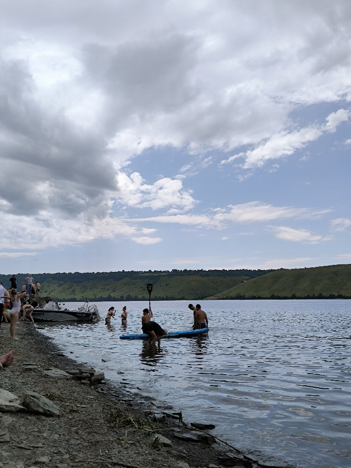 This image captures a daytime scene on the Dniester River near Horaivka, Ukraine. The sky is partially overcast, with dense grey clouds dominating the left and upper-center, transitioning to a clear blue sky with scattered white clouds on the right. The foreground consists of a rocky and gravelly shoreline that gradually slopes into the water.
Approximately 15-20 individuals are present in the scene. On the left side of the shoreline, a group of people are gathered, some standing and others partially obscured or seated on the rocks. A grey and black motorboat, bearing the visible registration "UMS-470", is partially beached on the left bank, adjacent to this group.
In the mid-ground water, at least seven individuals are visible. Centrally, two shirtless males are standing waist-deep next to a blue stand-up paddleboard (SUP); one of them holds a black paddle, while another individual is partially submerged and interacting with the SUP. To their left, a female in a dark two-piece swimsuit and another male are standing in the water. Other individuals are further back in the water, less distinctly visible. The water itself is calm with minor ripples.
The background is dominated by a continuous stretch of green, undulating hills covered in vegetation, extending across the horizon and bordering the wide river.