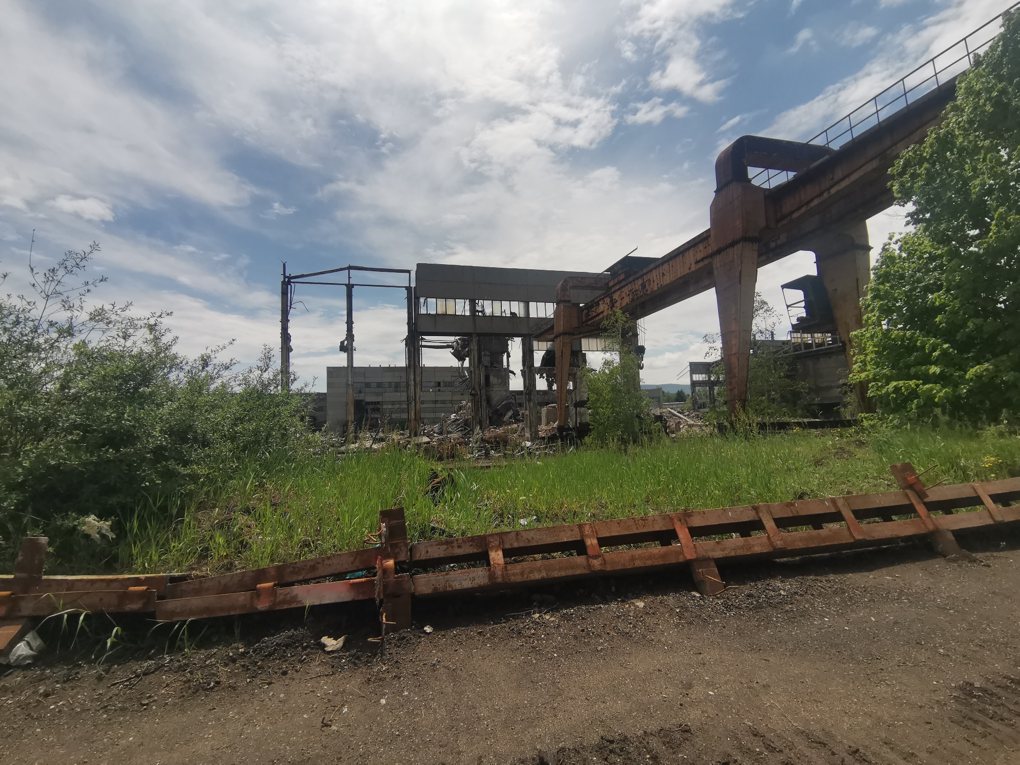 The image shows a derelict industrial site under a partly cloudy sky. The site appears to be in an advanced state of disrepair, with only skeletal remains of buildings visible. A large, rusty overhead crane dominates the right side of the frame. Debris litters the foreground, along with a rusty metal barrier that is lying on the ground. Vegetation is growing uncontrolled, further suggesting abandonment. The scene is located in the city of Verinsko, Bulgaria.