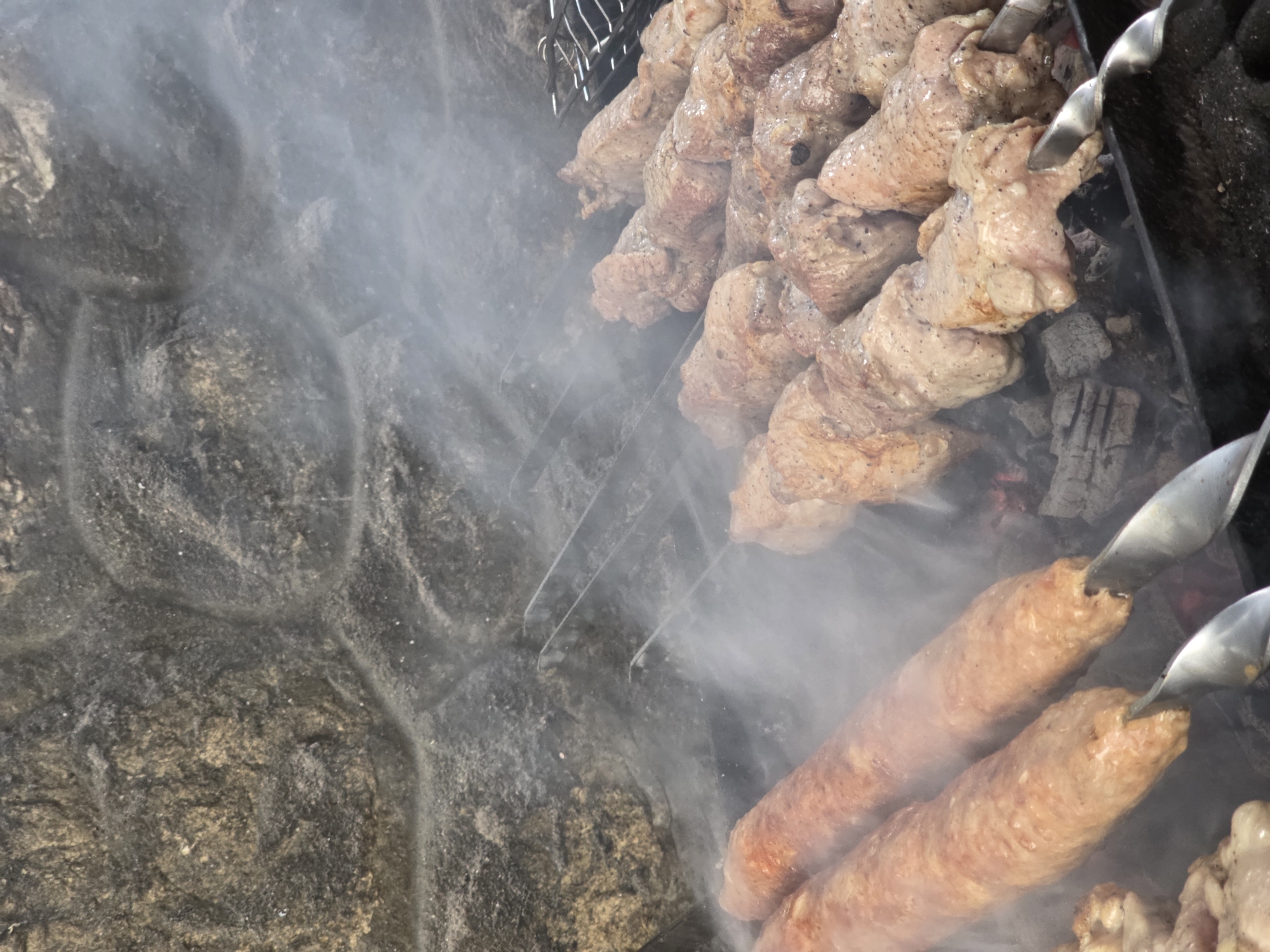 This media file shows a close-up of skewers of meat being cooked over hot coals, with smoke rising. The scene appears to be outdoors, with a rough, textured surface to the left of the food, possibly a stone wall. The lighting suggests it is daytime.
The primary subjects are skewers of meat. Some skewers hold chunks of meat, likely pork or beef, which are seasoned and appear to be in the process of grilling. Other skewers hold elongated, sausage-like meat, which are also cooking. The meat is a light brown color, indicating it is cooked or cooking. There are visible signs of charring on some pieces. The cooking apparatus includes metal skewers and what appears to be a metal grill grate. Hot coals are visible beneath the meat, providing the heat source, and smoke is billowing around the food, creating a hazy effect.
The activity is clearly the cooking of meat on skewers, a common method for preparing kebabs or shashlik. The event is likely a barbecue or outdoor meal preparation.
Notable details include the texture of the stone-like surface to the left, which is rough and uneven, with hints of tan and dark grey. The smoke is thick and obscures some of the details of the grilling area, adding to the atmosphere of cooking. The metallic sheen of the skewers is also noticeable. No people are visible in the media file. There is no visible text. The location context provided is Pidhorodne, Ukraine, suggesting a rural or suburban setting where outdoor cooking might be common.