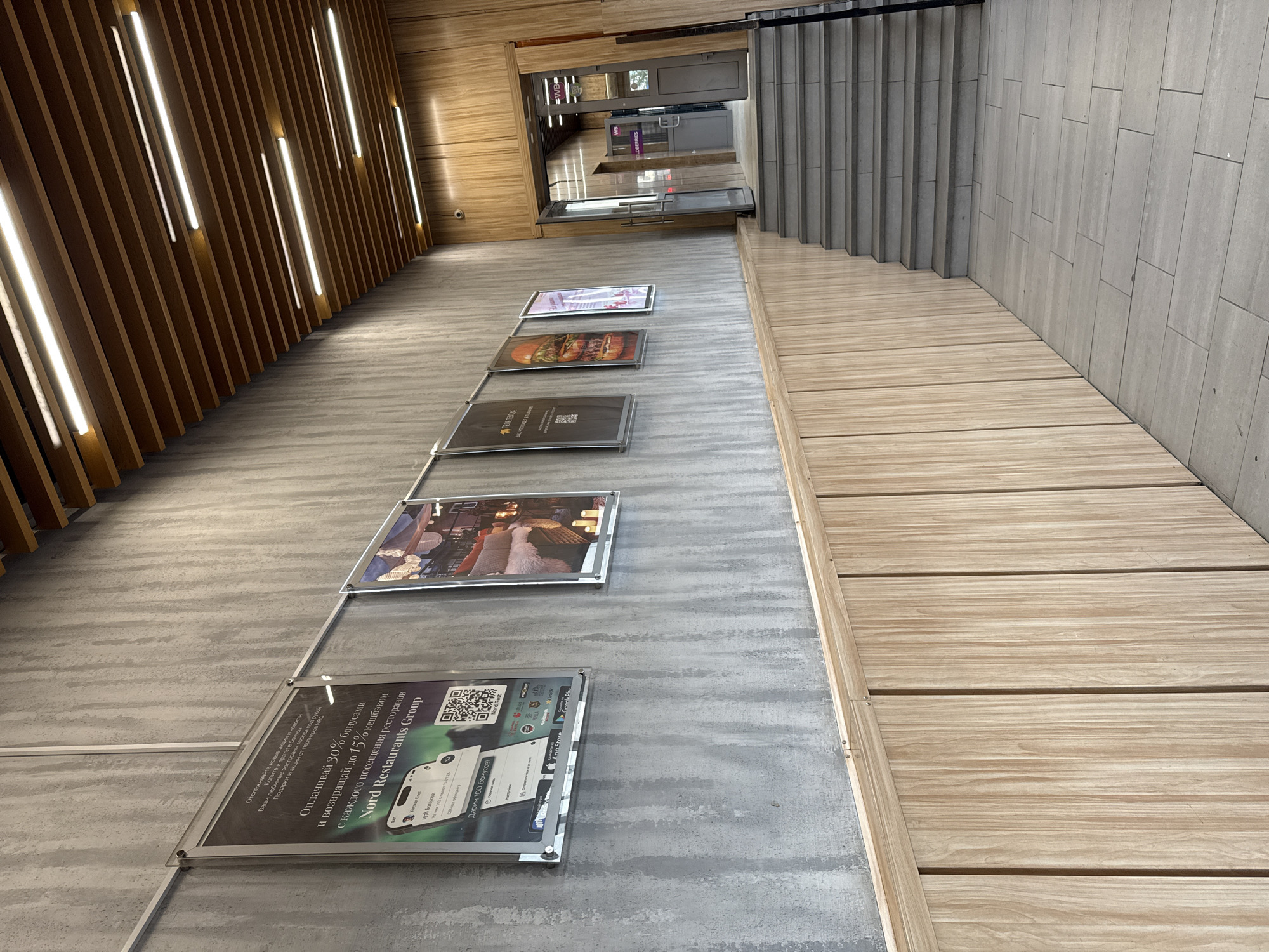 The image depicts the interior of a modern establishment, likely a restaurant or a shopping area, with a distinct architectural style. The walls are a mix of textured concrete and light-colored wood paneling, creating a contemporary and warm ambiance. A series of framed posters are displayed on the concrete wall, featuring advertisements and promotions for "Nord Restaurants Group" and other businesses, including an image of a burger.
The ceiling is a striking design element, constructed from parallel wooden beams with embedded linear LED lights, illuminating the space from above. A set of grey stone stairs ascends to the right, leading towards an open doorway. This doorway reveals more of the interior, including additional signage and what appears to be a retail space or another part of the establishment, possibly indicating a "WBB" logo. A security camera is visible on the wooden paneled wall near the ceiling.
The overall impression is one of a clean, well-lit, and contemporary commercial space. There are no people visible in the scene. Given the Cyrillic text on the advertisements, the location is likely in a Russian-speaking country. The text "Nord Restaurants Group" could suggest a business operating in the northern regions. Without further context or explicit signage within the image that directly mentions Norilsk, Russia, it is not possible to definitively confirm its presence in that specific city.
