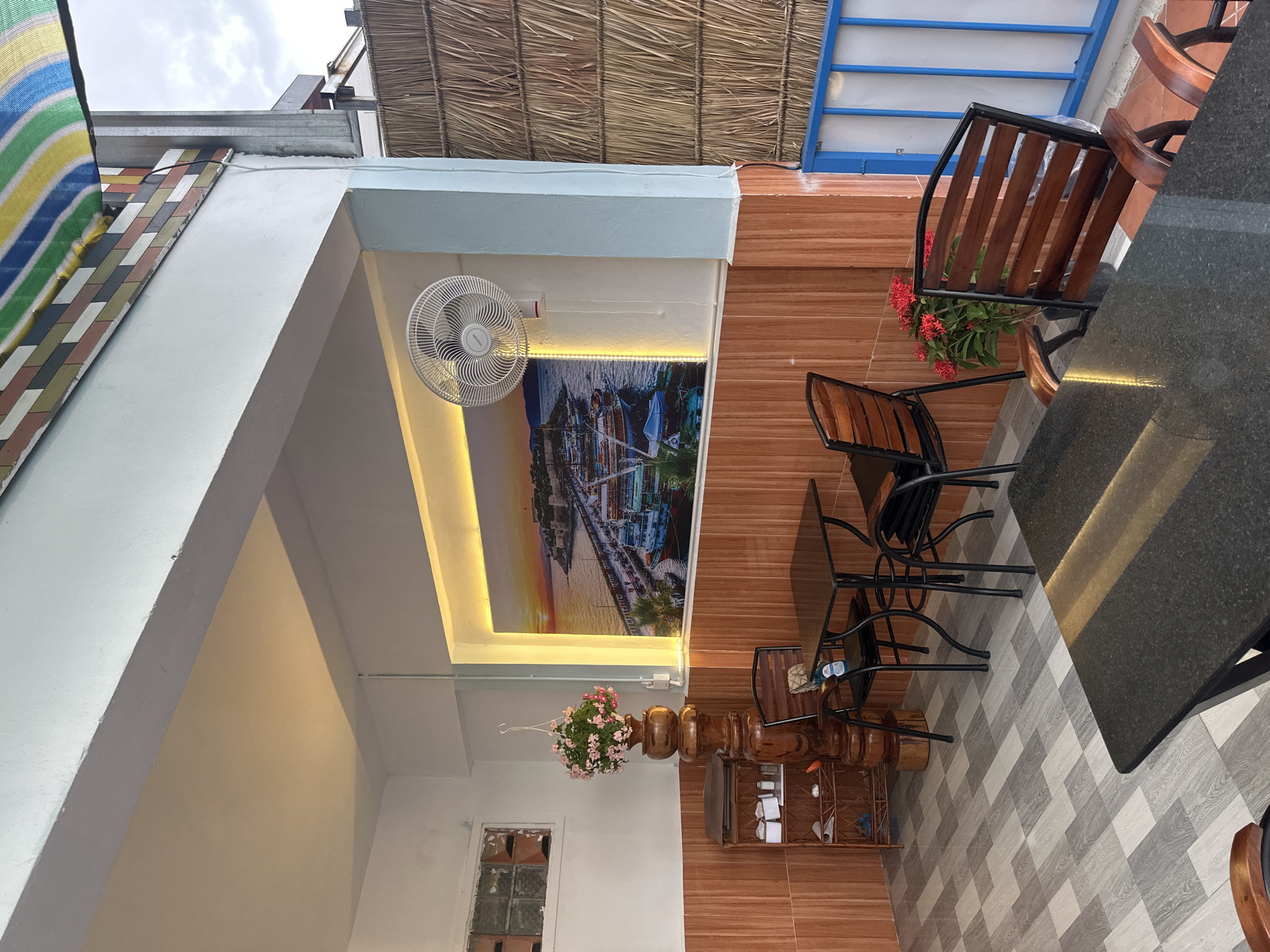 This is a ground-level, eye-level, medium shot of an outdoor seating area, likely a cafe or restaurant, in Kampot, Cambodia. The scene is captured during the day, with diffused natural light illuminating the space.
The seating area is covered by a roof, with a portion of a colorful, striped awning visible at the top. The walls are primarily clad in reddish-brown tiles, with a section of light blue paint on the right. A large, scenic mural of a harbor at sunset with boats and buildings is mounted on the back wall, illuminated by a yellow light strip. A white oscillating fan is mounted on the wall above the mural.
In the foreground, a dark, rectangular table with a glossy surface is positioned. To the left of this, a dark wooden chair with a slatted back and wooden armrests is visible, facing away from the camera. Further back, to the left, is a small wooden shelf unit holding various items, including white boxes. To the right of the shelf unit is a tall, ornate wooden pedestal supporting a hanging basket filled with pink flowers.
To the right of the pedestal, a square dark table is set with two black chairs. One chair, with a slatted wooden back and black metal frame, is pulled up to the table. Another identical chair is positioned to the right of the table. A small potted plant with vibrant red flowers sits on the floor near this table and chairs.
The floor is tiled in a geometric pattern of grey and light-colored rectangular tiles. A blue metal railing and a thatched wall are visible on the far right side of the image. The overall impression is of a relaxed, somewhat tropical outdoor dining space. There are no people visible in the frame.
