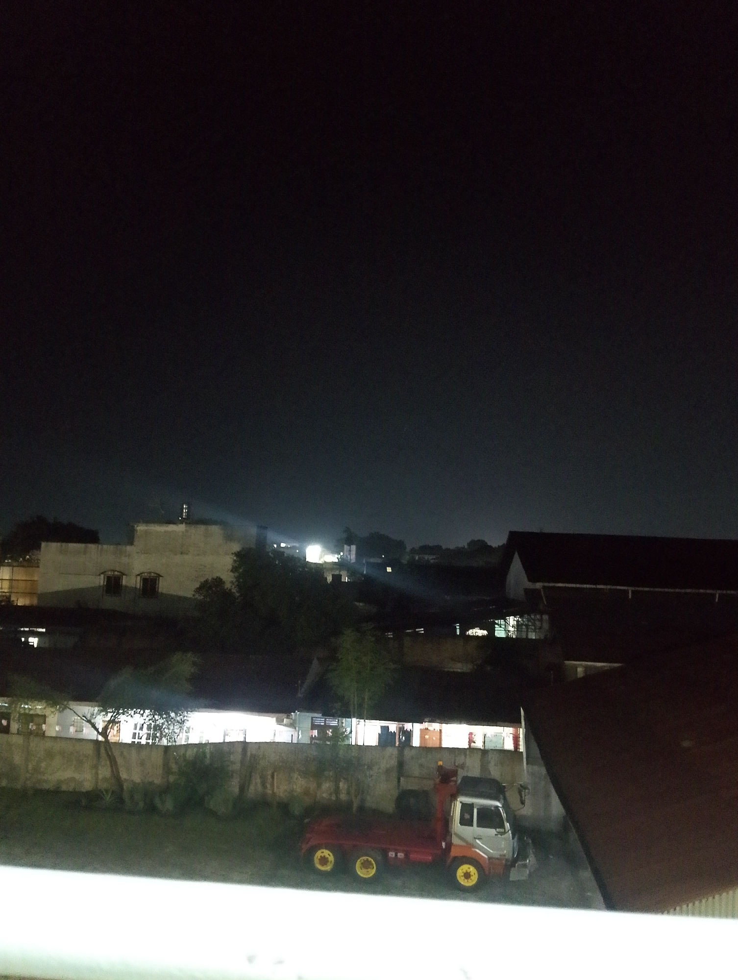The image depicts a nighttime urban scene in the City of Pematang Siantar, Indonesia, with a focus on residential and industrial structures under a dark sky. In the foreground, a small red and white truck with yellow wheels is parked in what appears to be an outdoor area. Behind the truck, a low concrete wall runs horizontally across the frame, separating the immediate foreground from a row of buildings further back. These buildings have illuminated windows, suggesting indoor activity. To the left, a lighter-colored building with a flat roof and visible windows is prominent. In the background, various other buildings and structures are visible, some with lights on, and trees dot the landscape. The overall impression is one of a quiet, late-night urban environment.