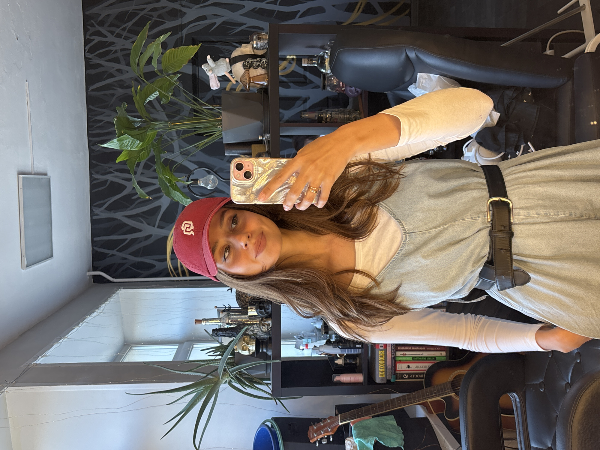 A young woman is taking a selfie in an indoor environment. She is wearing a maroon baseball cap with a white symbol on it, a white long-sleeved shirt, and a light blue denim dress cinched with a black belt. She is holding a smartphone with a patterned case.
Behind her is a bookshelf with various items, including books in Russian, a guitar, decorative objects, and plants. The background features a black and white patterned wall covering. A window is visible in the background, suggesting daylight. There is no indication of the city or location of Krasnoyarsk, Russia.