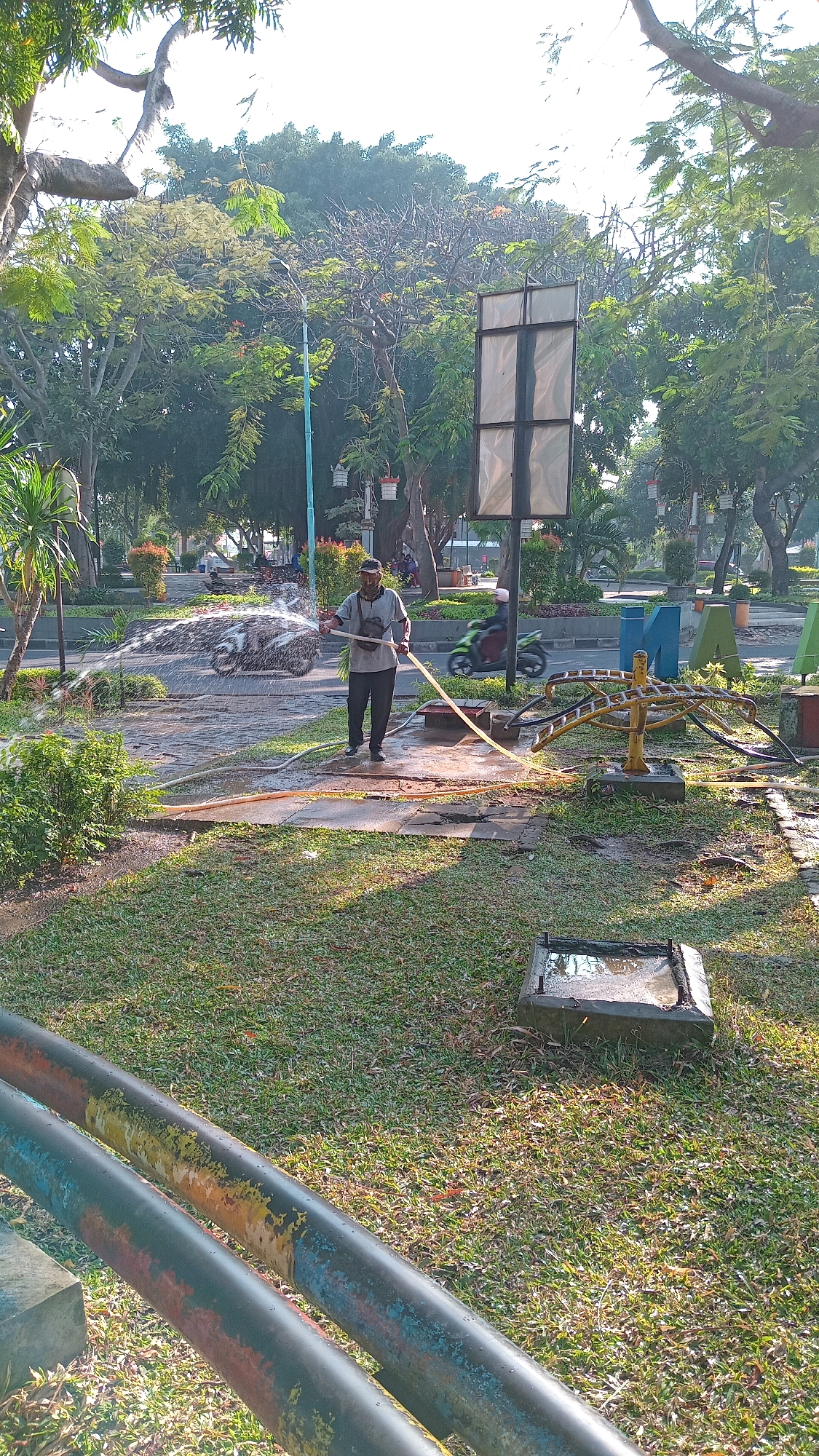 In Randuagung, Indonesia, a man is watering a grassy area with a hose in a park. He is wearing a dark cap, a light-colored t-shirt, and dark pants. He is holding the hose with his right hand and has a strap across his chest. The hose is spraying water onto the pavement and grass. In the background, there are trees, a road with passing motorcycles, and large decorative letters forming the word "MANDIRI". A signpost stands to the right of the man. The ground is wet from the watering. Two large, painted pipes are visible in the foreground, running diagonally across the frame.