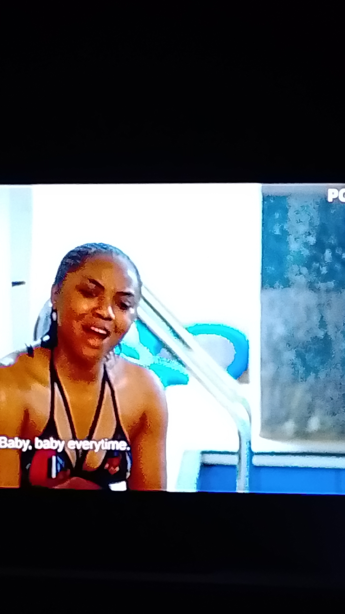 A woman with braided hair is sitting in a swimming pool and looking to the left with her mouth open as if she is singing. She is wearing a bikini top with a patterned design. There are blue pool stairs behind her. The text "Baby, baby everytime." appears on the screen in white lettering. The location in Lokoja, Nigeria is not specified.