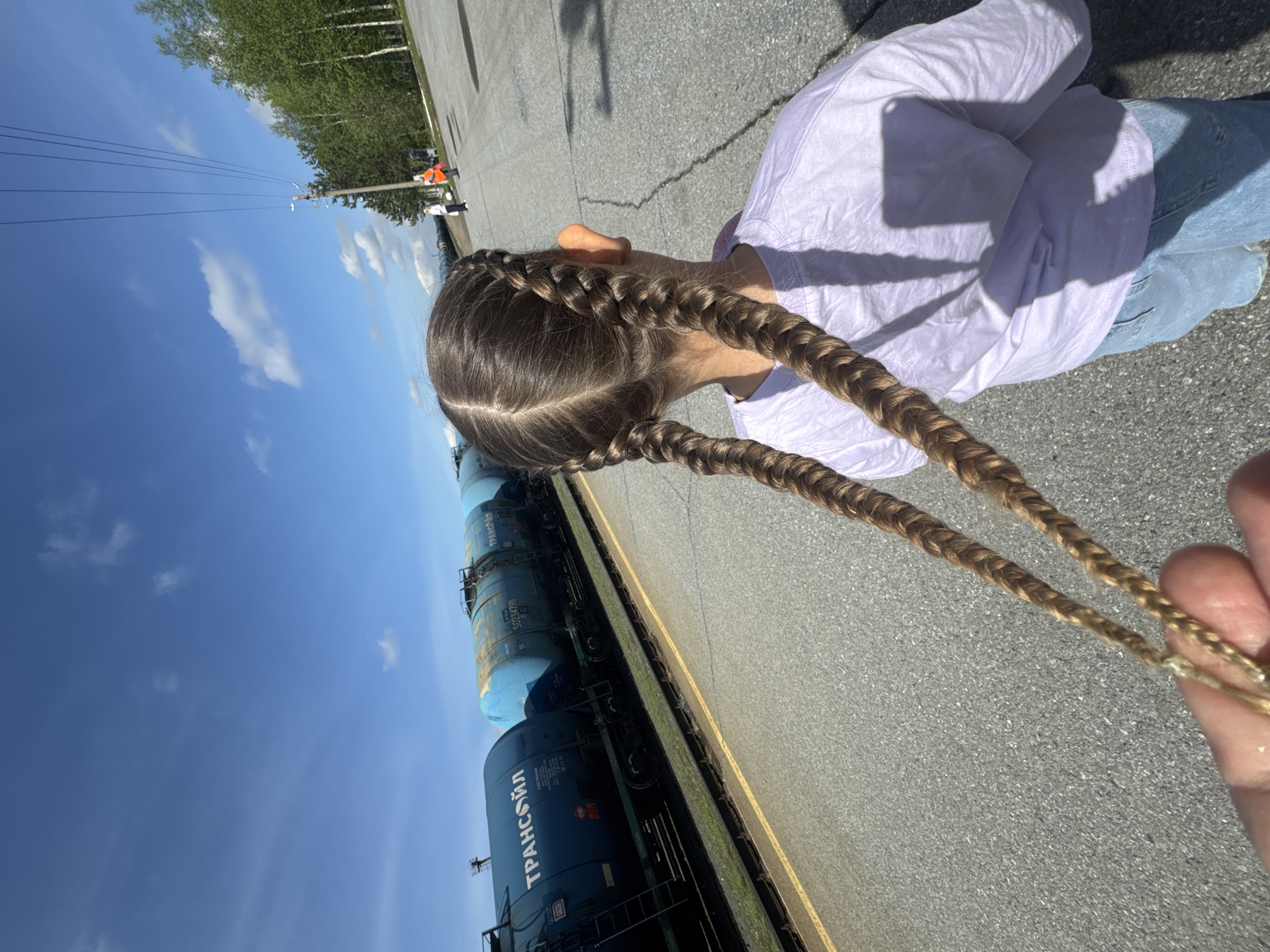 The image, set in Vysoky, Russia, depicts an outdoor scene on a bright, sunny day with a clear blue sky and scattered white clouds. The primary subject is a young person, likely a girl, seen from behind, wearing a light-colored long-sleeved shirt and light blue jeans. Her hair is styled in two long, prominent French braids that extend down her back. A hand in the lower right corner casually holds one of these braids.
The person stands on an asphalt surface marked with a yellow line, positioned next to a railway track. Along the track, several train tank cars are visible. These cars are dark blue/black and light blue, and feature Cyrillic text. Visible text includes "ТРАНСОЙЛ" (TRANSOIL) and identification numbers such as "50234778" and "50239109" on the darker cars.
In the background, beyond the railway line, a line of green trees indicates a natural or semi-natural setting. A utility pole with wires is also visible in the distance, and faint figures, one wearing an orange vest, can be discerned near it. The bright daylight casts distinct shadows, including a clear shadow of a smartphone on the subject's back and leg, suggesting the photo was taken from above. The scene captures a casual moment by a railway.
