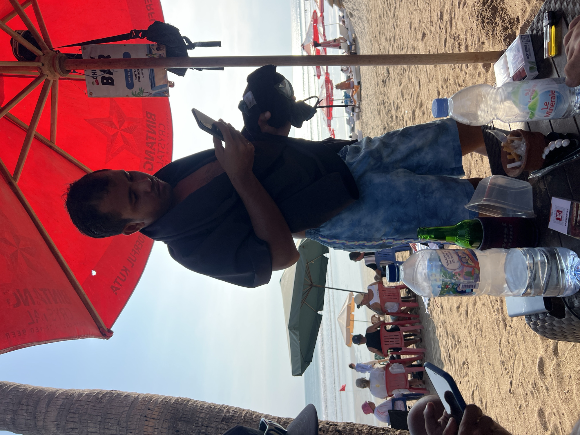 A man stands on a sandy beach in Kuta, Indonesia, holding a mobile phone in his right hand and a black fabric item in his left. He is wearing a dark t-shirt and blue patterned shorts. In the foreground, a table holds two bottles of water, one branded "Le Minerale," a green bottle, a cigarette pack, and a lighter. Behind the man, beach umbrellas and chairs are visible, along with other beachgoers. The ocean and sky form the backdrop. The scene appears to be a typical day at a beachside establishment.