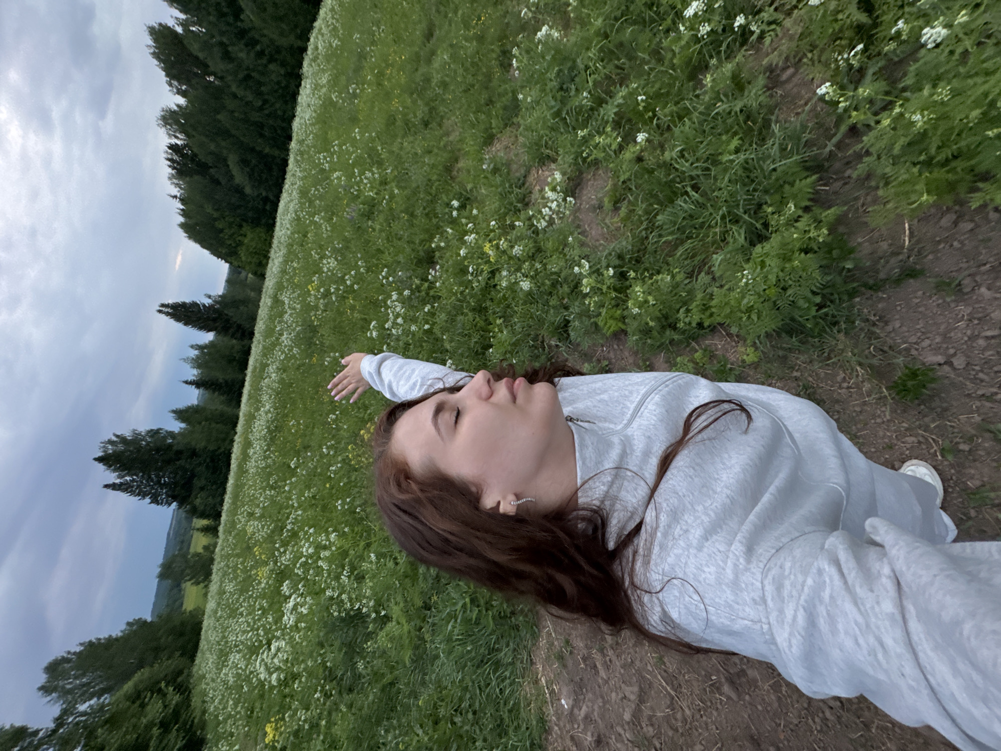 A young woman is lying in a field in Nizhnyaya Gar', Russia. She has brown hair and is wearing a gray sweatshirt and white sneakers. She is lying on a dirt path with a field of green grass and small white flowers to her left. There are trees in the background and a cloudy sky overhead. She has her right arm raised.