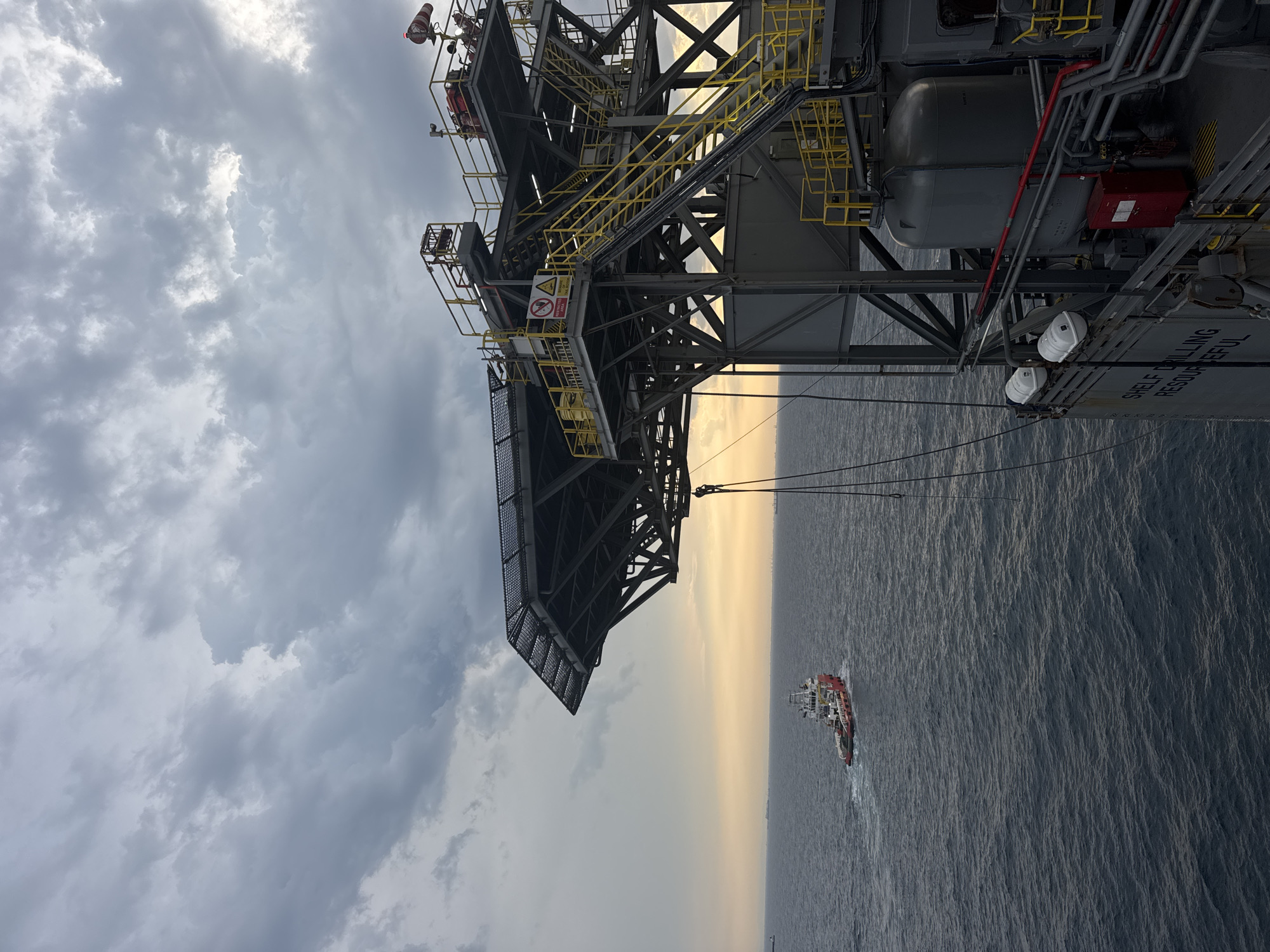 The image captures an offshore oil rig structure against a cloudy sky and a calm sea. A tugboat is visible in the distance, navigating the water. The rig features metal walkways, staircases, and industrial equipment. The scene appears to be at dusk or dawn, indicated by the soft, warm glow of the sun near the horizon. There are no people visible in the image. The location is not in a city, but rather offshore.
