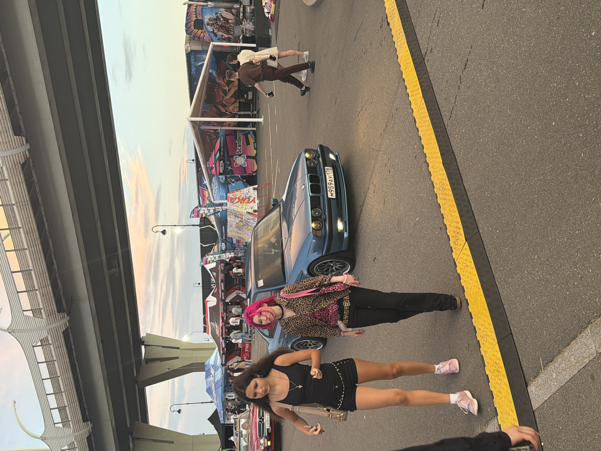 Two young women walk on asphalt near a blue BMW. The woman on the left wears a black tank top and black shorts, with a beige purse slung over her shoulder. She has dark brown hair and is wearing pink sneakers. The woman on the right has bright pink hair and wears a leopard print shirt, black pants, and black shoes. She carries a pink purse. In the background, tents and a large truck with graffiti are visible. A large concrete overpass looms overhead. The scene appears to be an outdoor event.