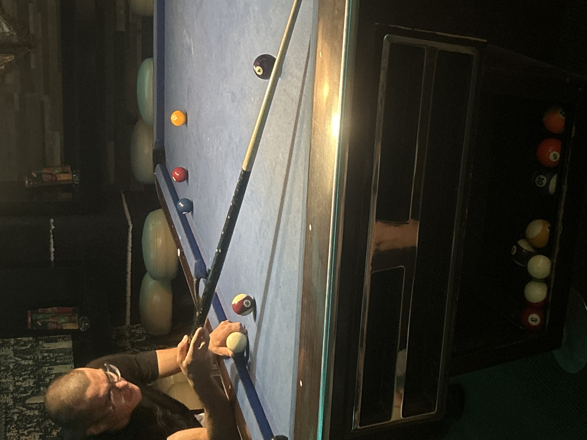 A man with glasses prepares to strike a cue ball on a blue felt pool table. Multiple pool balls are scattered on the table. Several balls are in the ball return area. The location cannot be determined based on the image, and there is no evidence it is in Pleven, Bulgaria.