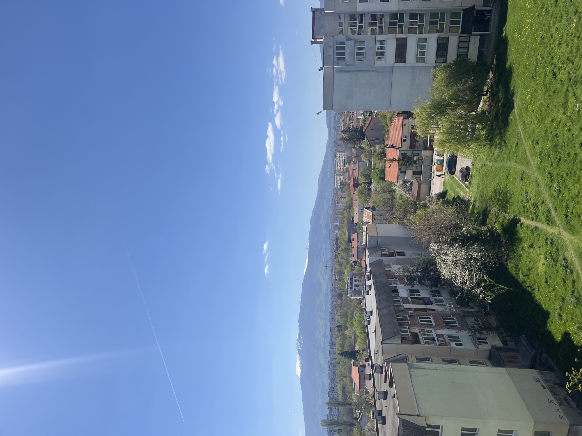 The image shows an urban landscape in what is likely the city of Sofia, Bulgaria, judging by architectural features and mountain range in the background. The scene is captured during daylight with a bright blue sky marked by thin contrails and a few scattered clouds.
The view is an elevated vantage point overlooking residential buildings with varied heights and designs, including apartments in muted colors and some with red-tiled roofs. Trees are interspersed amongst the buildings, with green foliage suggesting either springtime or summer. A mountain range is in the distance, showing hints of snow on its peaks.
On the right edge of the image is a green space. The presence of footpaths suggests it’s a public area, possibly a park or a landscaped area within a residential complex. The image does not contain people or actions.