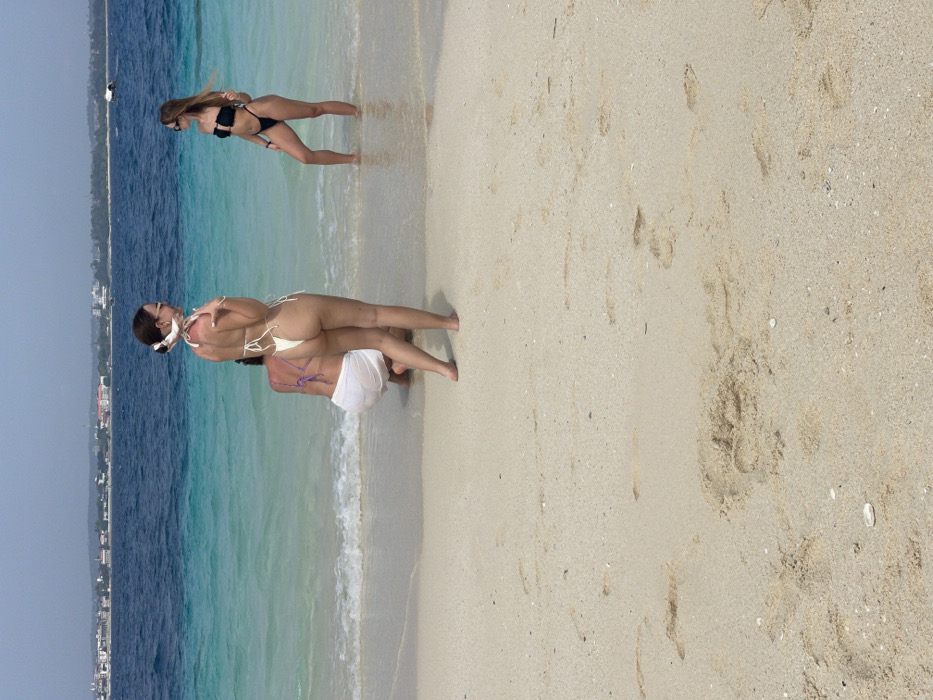 The image depicts three adult females on a sandy beach with clear, turquoise ocean water, facing the distant cityscape of Zanzibar, Tanzania. The sky is clear. In the foreground, the light-colored sand shows various footprints. Centrally positioned, a female stands with her back mostly to the camera, wearing a cream-colored string bikini and sunglasses, with her hair in a bun styled with a white scarf. To her left and slightly behind, another female kneels in the shallow water, wearing a purple bikini top and a translucent white sarong. To the right, a third female stands in the shallow water, facing left towards the others, wearing a black strapless bikini and sunglasses. The ocean transitions from light turquoise to a darker blue, with a discernible coastline and buildings in the distant background. A small white boat is visible on the water near the horizon.