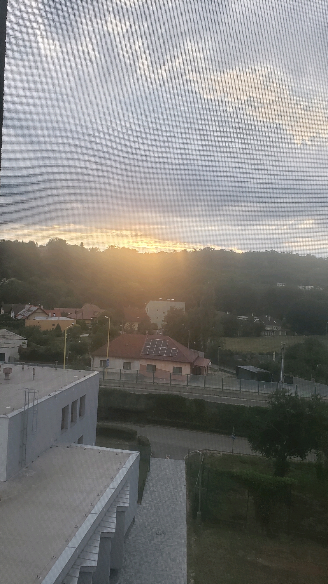 The image depicts a panoramic view of a residential area in Prešov, Slovakia, as seen through a window screen. The setting sun casts a warm glow over a landscape of rolling hills covered in dense foliage. Scattered among the trees are several houses with red-tiled roofs. One house in the foreground prominently features solar panels. In the middle ground, a street runs parallel to the houses, with a few streetlights visible. To the left of the frame, a modern, light-colored building dominates the foreground, with a grey tiled roof and several windows. The sky is overcast with streaks of sunlight breaking through, suggesting either dawn or dusk. The overall impression is one of a peaceful, semi-rural suburban environment.