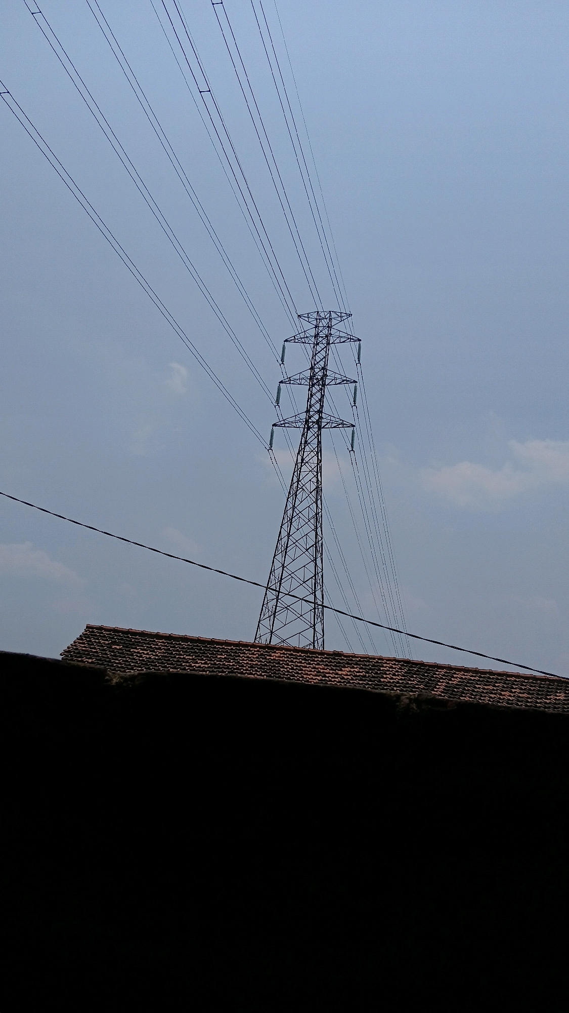 A high-angle, outdoor, full shot shows a pylon and power lines against a hazy sky. The pylon is a tall, latticed metal structure with thick cables suspended from it. The cables stretch across the frame from left to right, creating a diagonal line. Below the pylon is a tiled roof, partially obscured by a dark, blurry foreground object. The sky is overcast and a pale blue. The scene appears to be located in Klorogan, Indonesia, as indicated by the context. There are no people or visible actions in the image.
