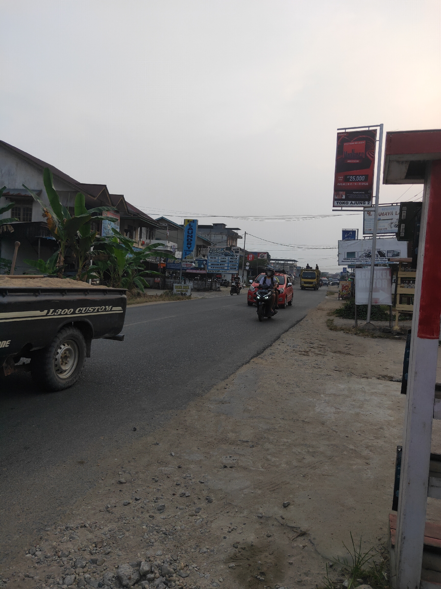 The image depicts a street scene in Sintang, Indonesia. A black pickup truck, labeled "L300 CUSTOM," is parked on the left side of the frame, loaded with sand. The truck's rear wheel and part of its body are visible. The road, a mix of asphalt and gravel, stretches into the distance.
Along the roadside, there are several buildings, including houses with brown roofs and some commercial establishments. One building displays a sign for "TOKO AJUNG 2" and a billboard advertising Marlboro cigarettes. Another sign reads "Roli EXPRESS LAUNDRY."
Several vehicles are present on the road. A red car is driving away from the camera, followed by a motorcycle with a rider wearing a helmet. Further down the road, a yellow truck is also visible. Other motorcycles and cars can be seen in the distance.
The sky is overcast with a soft, diffused light, suggesting it might be early morning or late afternoon. Banana trees and other vegetation are interspersed with the buildings, typical of the tropical climate. The foreground on the right shows a concrete pathway and some sparse grass. The overall impression is of a moderately busy street in a semi-urban area.