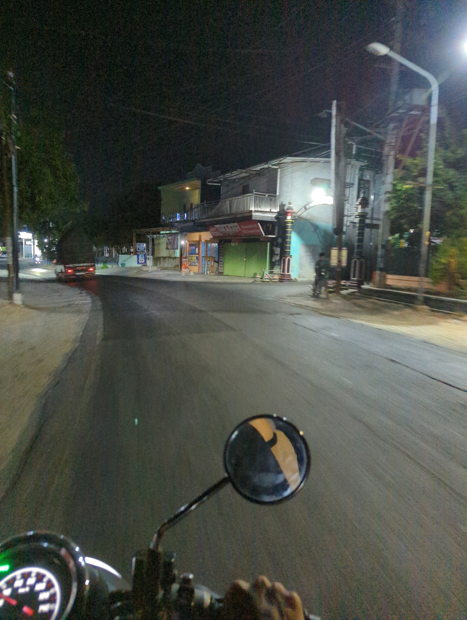 The image depicts a nighttime scene on a street in Kacangan, Indonesia. The perspective is from a moving vehicle, likely a motorcycle, with the dashboard and a rearview mirror visible in the foreground. The road surface is dark asphalt, illuminated by streetlights and the lights from buildings.
On the left side of the road, there is a sidewalk with a tree and a pole. Further down the road, a truck with its taillights illuminated is visible. On the right side of the road, there are several buildings with lights on, suggesting they are businesses or residences. A streetlamp is prominent on the right side of the image, and there are power lines and other utility poles. In the distance, a person is seen standing near a building. The overall atmosphere is one of a quiet, urban night.