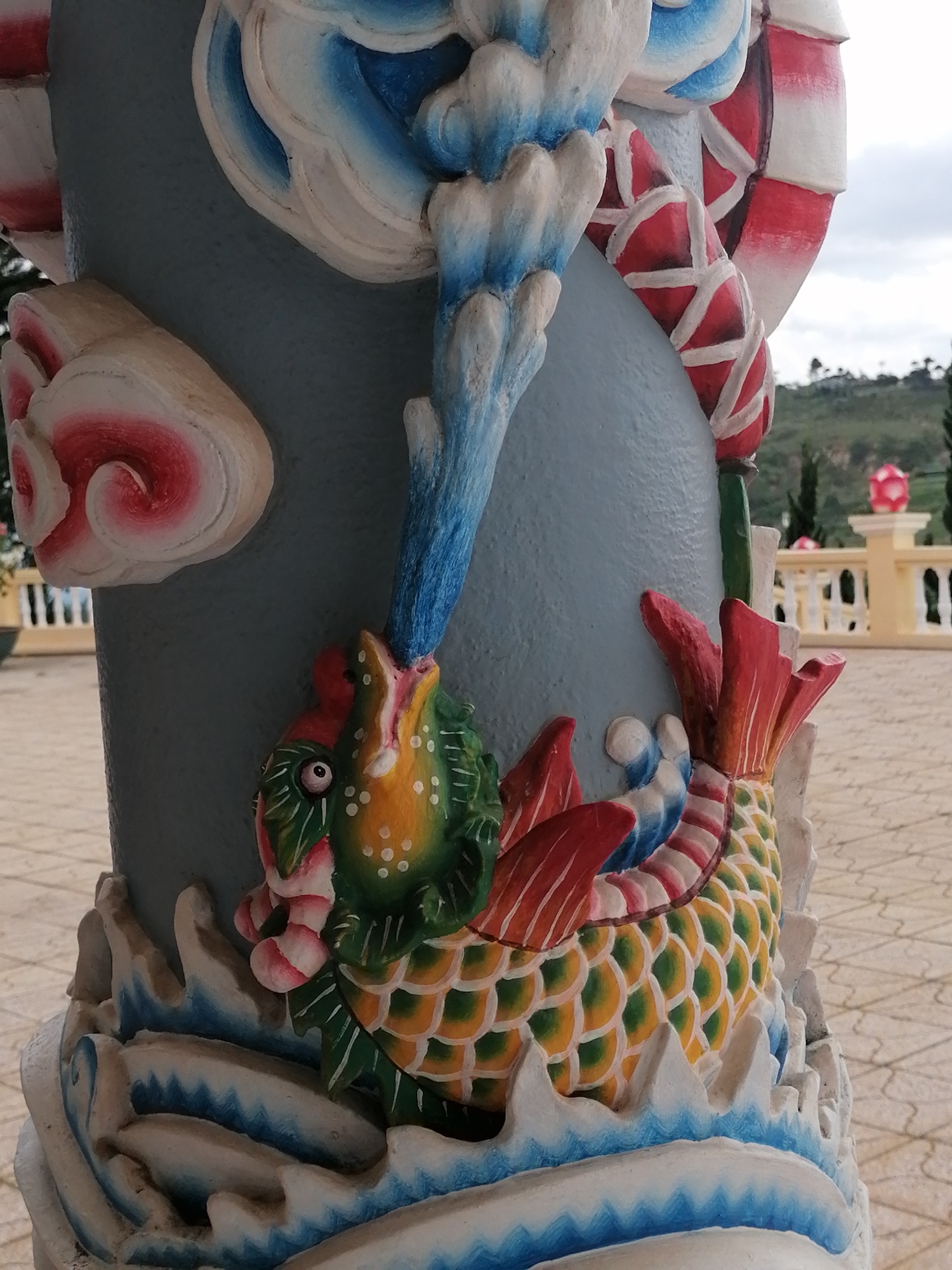The image shows a close-up of a decorative column, likely part of a temple or shrine, in Lộc Quý, Vietnam. The column is adorned with colorful bas-relief sculptures. In the foreground, a mythical creature, possibly a dragon or fish, is depicted with scales of green and yellow, a red tail fin, and a green head with a red and white striped mouth. Above the creature, blue and white cloud-like decorations are visible, along with red and white striped bands. The background shows a light-colored tiled ground, a white railing, and a rolling landscape with trees and a pink spherical object on a pedestal. The sky is overcast.