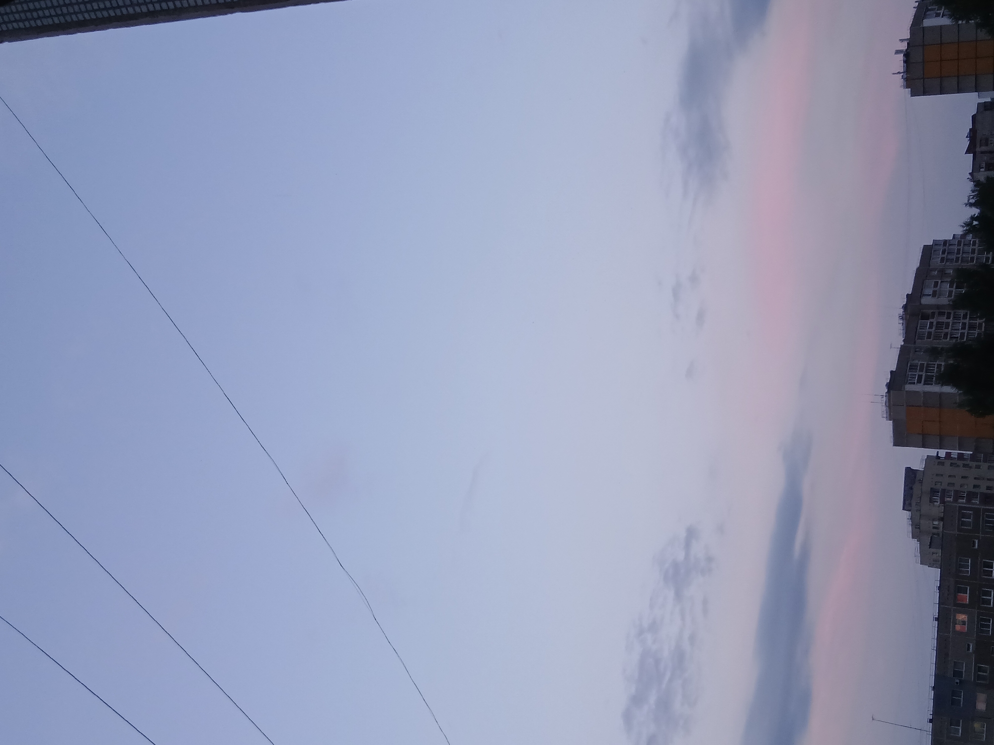 The image depicts a twilight sky with a pale blue and pink hue over a city skyline. Three thin wires are stretched across the upper portion of the frame. The lower part of the image shows the tops of several apartment buildings, some with lit windows. Trees are visible in front of the buildings. The overall scene suggests a residential urban environment at dusk.