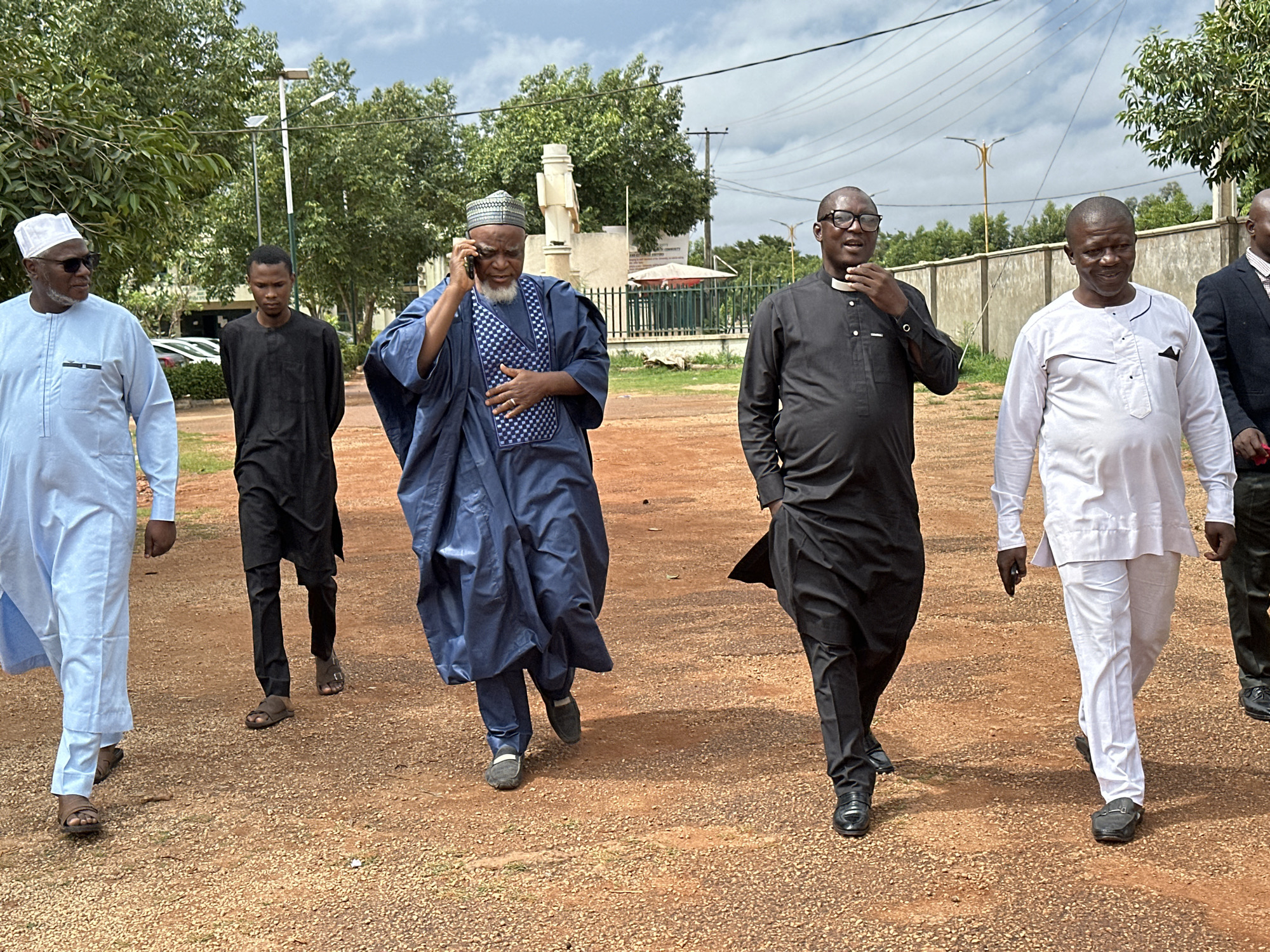 Six men are walking on a reddish-brown dirt path with gravel. The man on the far left is wearing a light blue traditional outfit and a white cap. Next to him is a young man in a black traditional outfit. The third man from the left is wearing a dark blue traditional outfit and a patterned cap, and he is holding a phone to his ear. The fourth man from the left is wearing a black traditional outfit and glasses, and he is touching his collar. The fifth man from the left is wearing a white traditional outfit. The sixth man from the left is wearing a dark suit and a red and white patterned shirt. Trees and utility poles are visible in the background, and the sky is cloudy. The location is identified as Kabala, Nigeria.