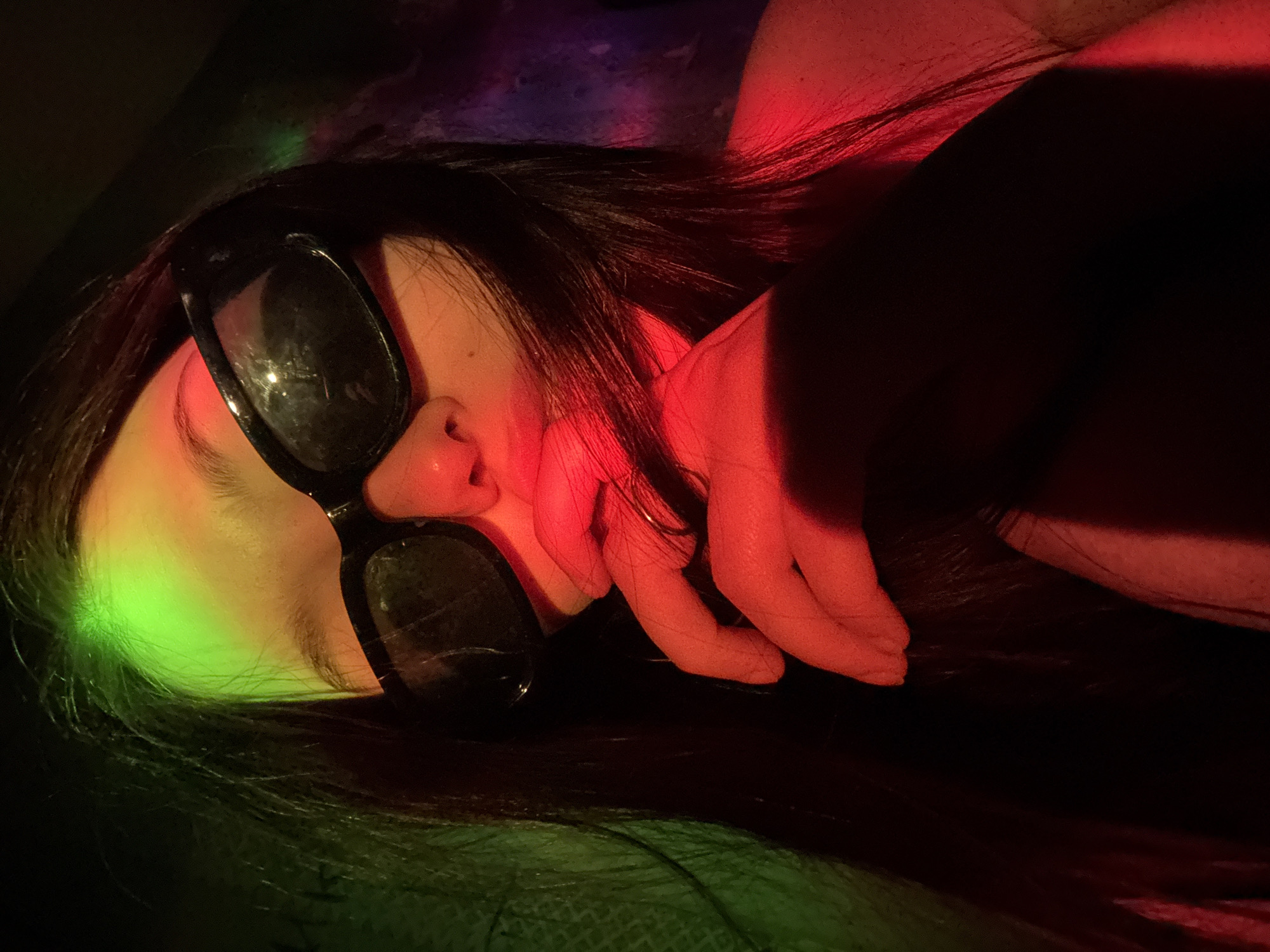 A person with long, dark hair lies on their side wearing sunglasses. Red and green light illuminate different sections of their face and hair. Their hand rests near their face. The background is dark and indistinct. The location in the city of Фоки, Russia is not evident in the image.