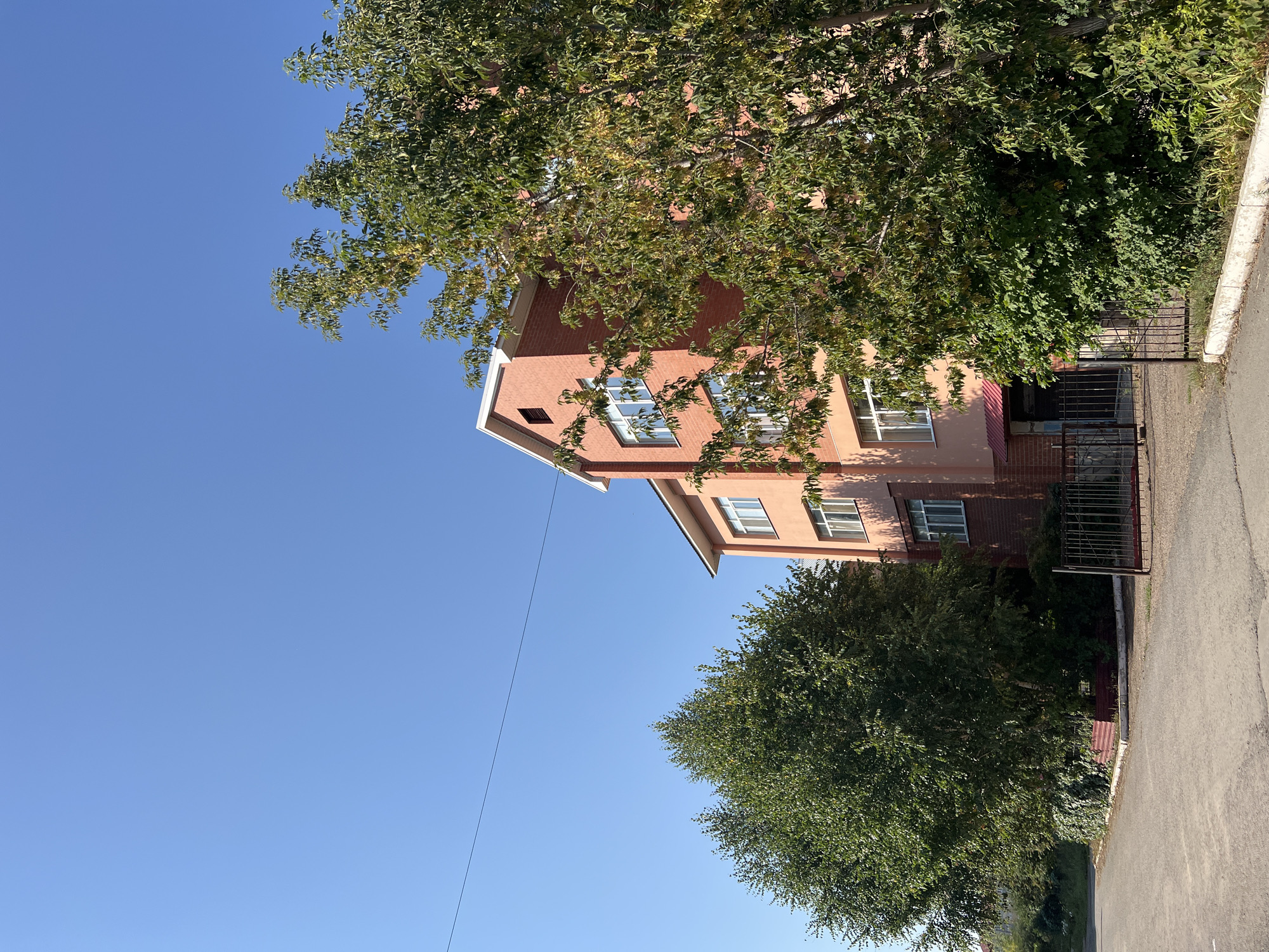 A multi-story building with orange and red brick facade is seen under a clear blue sky. The building has multiple windows with white frames and a red roof visible on the top floor. Trees with green foliage are in front of and to the side of the building. A black metal fence is in front of the building. The ground in front of the building is paved with asphalt. The scene is likely a residential or educational building located in the city of Salavat, Russia.