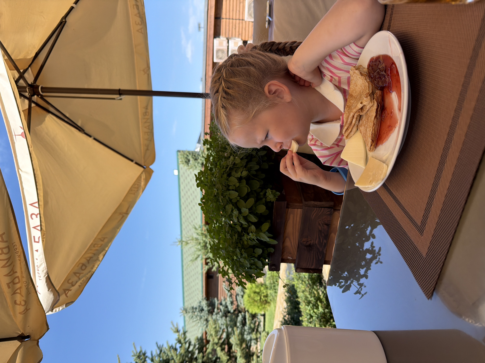 A young Caucasian girl with blonde hair styled in braids is seated at a table under a yellow umbrella, eating cheese. She is wearing a pink and white striped shirt. On the table in front of her is a white plate with pancakes, cheese slices, and jam. The background shows a courtyard with trees, a planter box with green foliage, and buildings with green roofs. The scene appears to be a cafe or restaurant in Kamyshin, Russia. The sky is clear and blue.