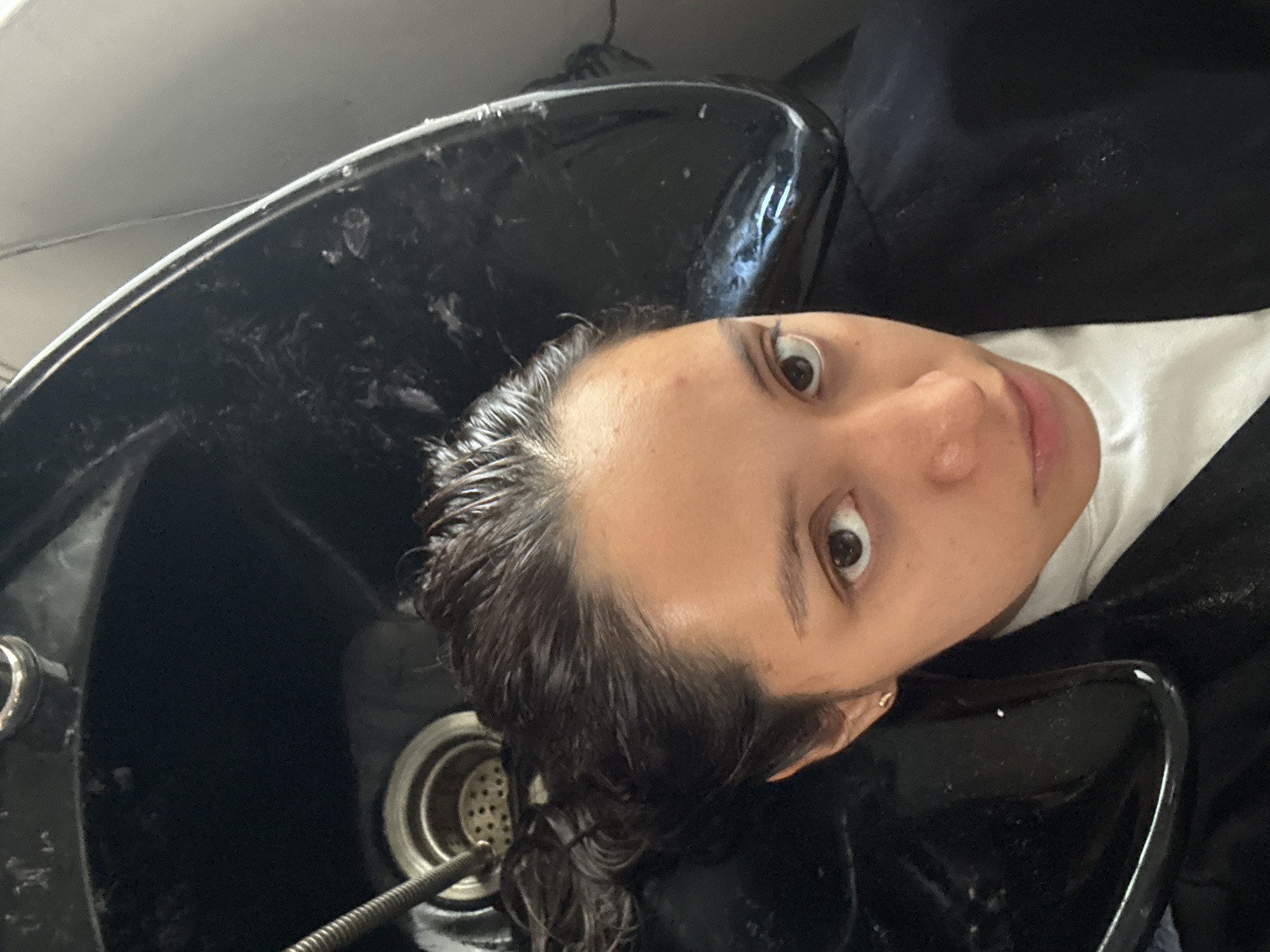A woman with wet, dark hair is reclined back into a black hair salon washing station. Her face is angled towards the camera, with wide eyes looking directly at the viewer. She is wearing a white t-shirt and a black salon cape. The washing station is also black, with a visible drain and hose. The location is a hair salon, presumed to be in Puebla, Mexico.
