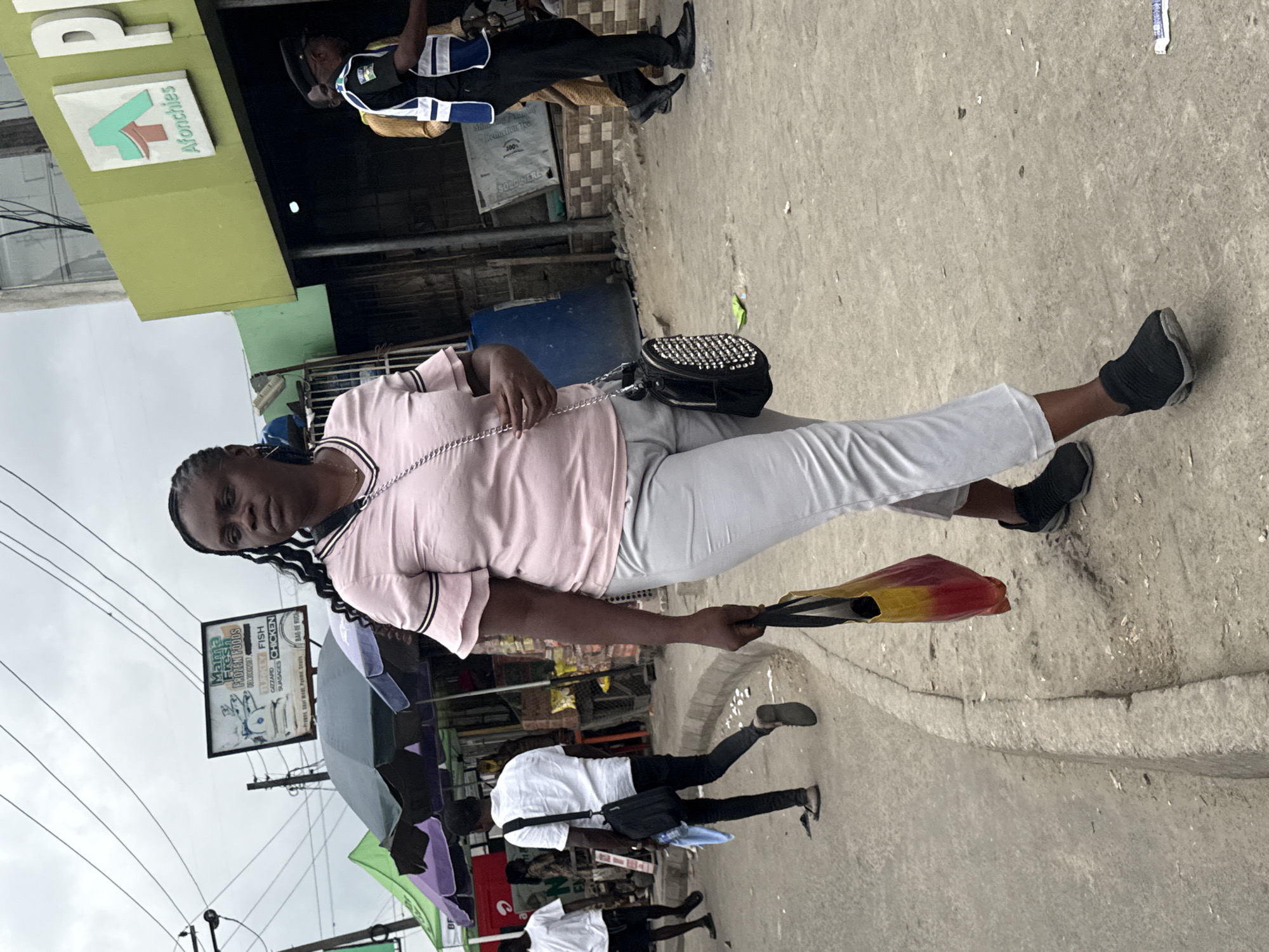 The image shows a street scene in Ado, Nigeria. A woman in a pink shirt and grey pants walks towards the viewer, carrying a red and yellow bag in her right hand and a black studded purse on her left shoulder. To her left, a man in a white shirt and black pants walks away from the viewer, carrying a black bag. Further in the background, a security guard in a blue and white vest stands near a building with a green facade. The environment appears to be a busy urban area with stalls and a cloudy sky.