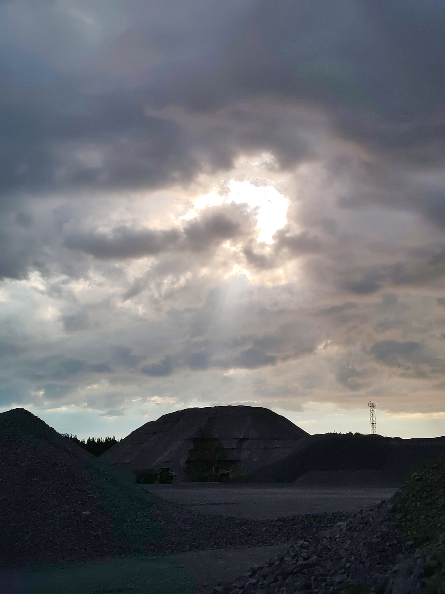 This is a medium-format, high-angle, vertical, color, exterior, daylight photograph of a gravel pit in Shalsky, Russia. The sky is filled with dark, heavy clouds that are partially broken in the center, allowing a bright shaft of sunlight to penetrate. In the foreground, on the left, is a large pile of dark gray gravel. To the right of that, a flat, gray gravel area extends back to a large, dark, conical pile of material, possibly coal or slag. In front of the large pile, two dark, heavy machinery vehicles are visible. Further to the right, another, smaller pile of dark material is visible, with a distant, lattice-work communication tower with red and white markings standing against the sky. The scene conveys a sense of an industrial or mining environment under a dramatic sky.