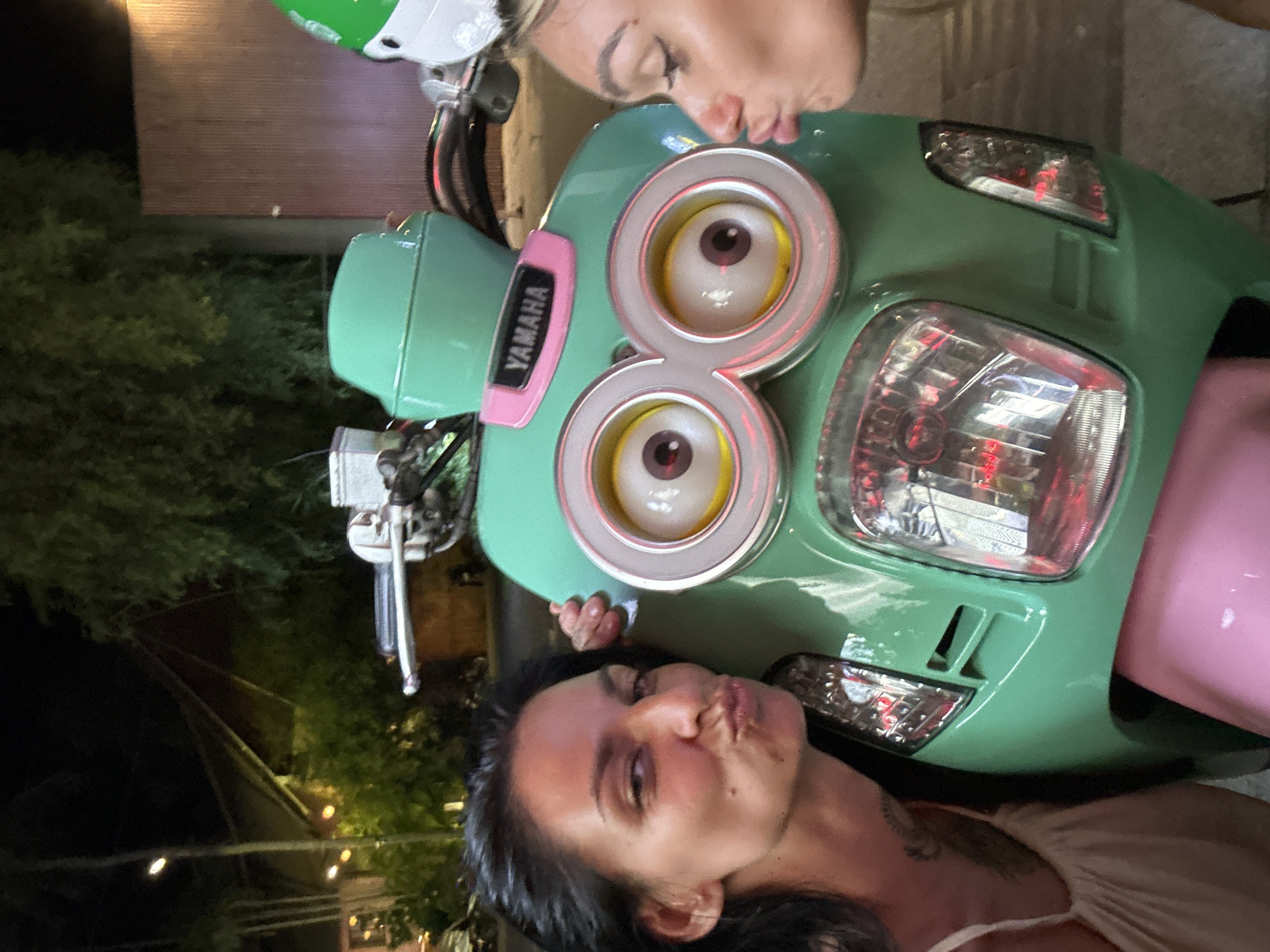 Two women are posing with a green Yamaha scooter that has been decorated to resemble a Minion character, with two large circular eyes. The woman on the left, with dark hair, is leaning towards the scooter and puckering her lips, with her right arm resting on the scooter. The woman on the right, with blonde hair, is also leaning towards the scooter and puckering her lips, with her face partially obscured by her hair and the scooter. The scooter is parked on a sidewalk, with a building and trees visible in the background. The scene appears to be in Phu Thuy Ward, Vietnam, as indicated by the scooter's branding and the tropical foliage.
