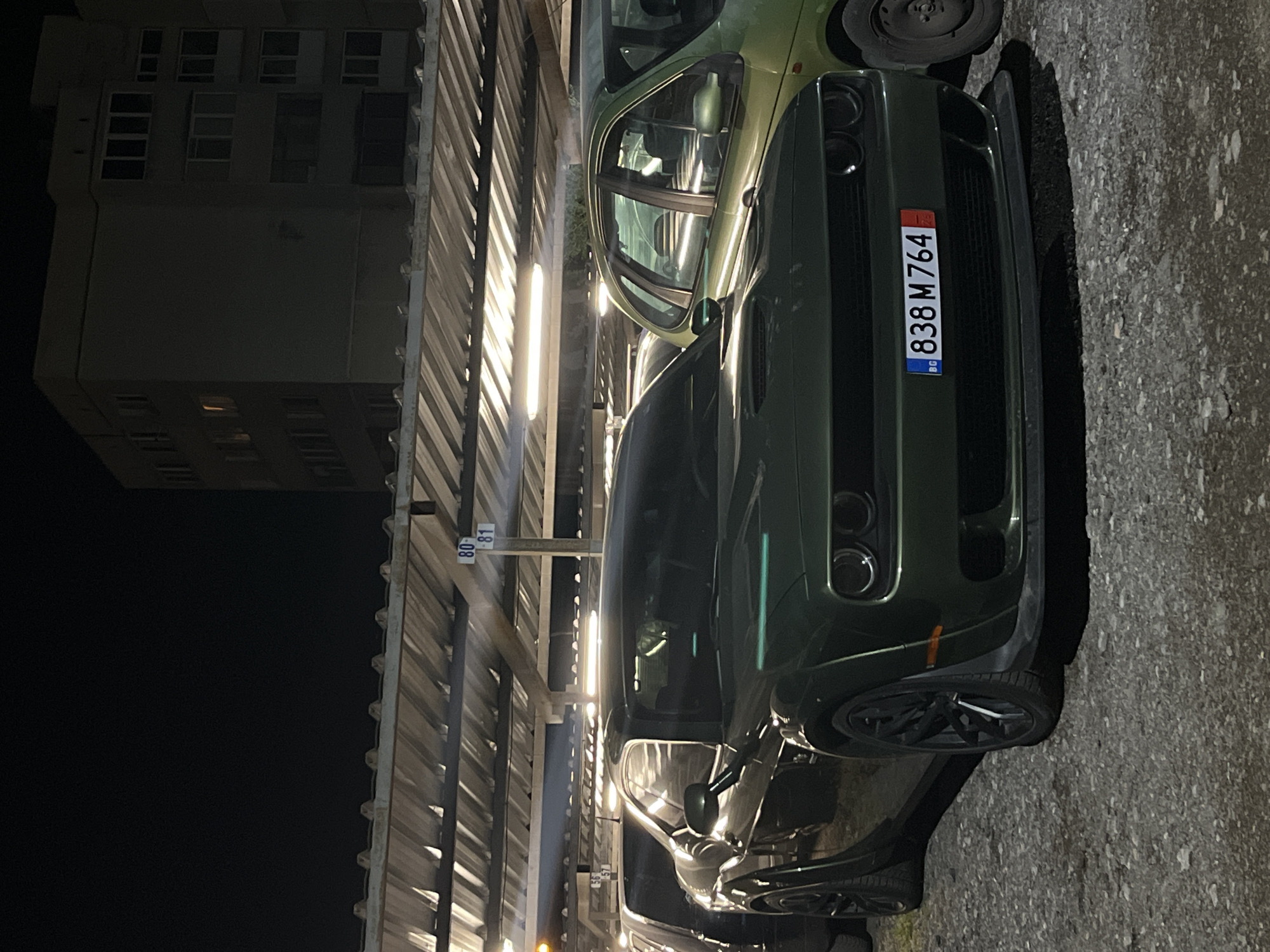 The image shows a dark green Dodge Challenger parked in a parking structure in Sofia, Bulgaria. The license plate number is 838 M 764. The background includes a tall building and the structure of the parking facility. The lighting suggests it is nighttime. Another car, possibly a green Opel, is parked behind the Dodge. The location appears to be a parking lot possibly connected to a residential or commercial building.