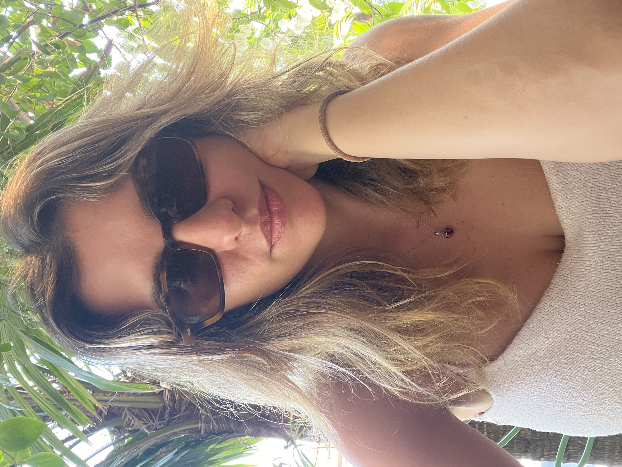 The image is a selfie of a woman with blonde hair and sunglasses, resting her chin on her hand. She is wearing a white top and a necklace with a red pendant. The background shows palm trees and greenery, suggesting an outdoor tropical setting. The lighting indicates it is daytime. The woman appears relaxed and is looking directly at the camera. The location is identified as Mengwi, Indonesia.