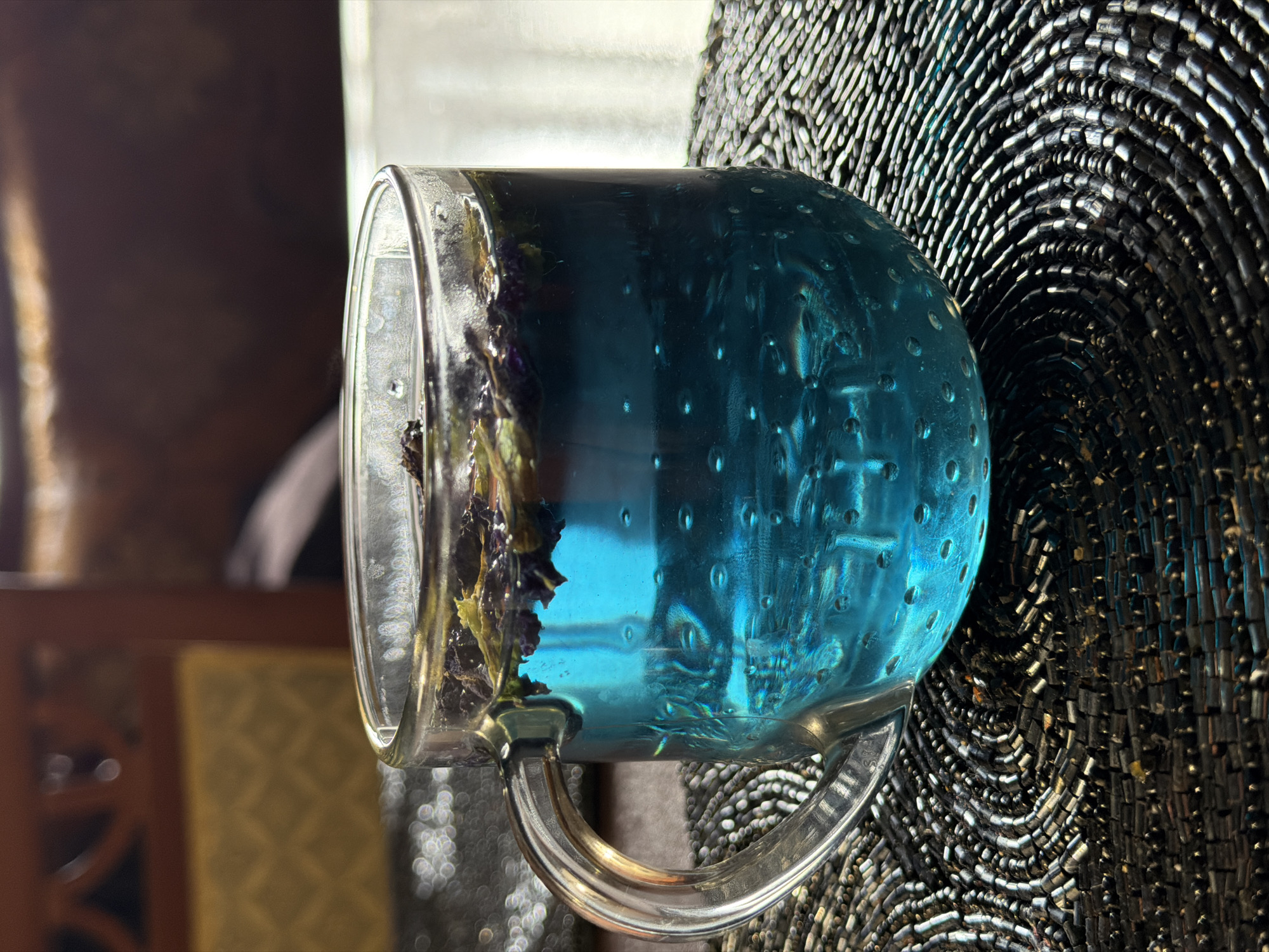 A clear glass mug filled with bright blue butterfly pea flower tea sits on a dark, beaded placemat. The tea has some dried flower petals floating on the surface. The mug has a handle and the glass is slightly textured, creating small bubbles within the liquid. In the background, out of focus, is a wooden piece of furniture, possibly a chair or cabinet, with a textured, gold-colored upholstered section. The lighting suggests an indoor setting. The scene does not provide any specific indicators of its location within the city of Surgut, Russia.
