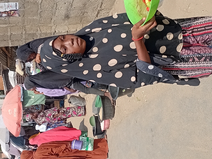 A woman wearing a black polka dot headscarf and colorful skirt holds a green bowl with red and green items. She is outdoors with other people and vendors' stands in the background. Some stands have umbrellas. The ground is light gray.