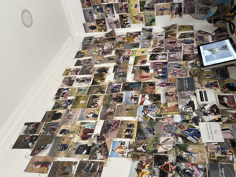 The image depicts an indoor room in Çiftlikköy, Turkey, with white walls almost entirely covered by a dense, overlapping collage of hundreds of printed photographs. These photographs, generally small to medium in size, primarily feature numerous individuals and groups, predominantly young adults, participating in various outdoor activities. A significant recurring theme is the presence of All-Terrain Vehicles (ATVs) or quad bikes, with subjects shown riding, posing alongside, or standing within natural, often hilly, landscapes. Many of the prints display curled edges and wear. Visible text on some photographs includes Arabic script and English phrases such as "WE HAVE CURRENCY EXCHANGE" and "One into Good". In the bottom right, a desk holds an open silver laptop displaying an image on its screen and a blue-bladed electric fan with a yellow base. A clear plastic water bottle with a blue cap is partially visible on the desk. The white ceiling features a single circular recessed light fixture. The walls exhibit plaster imperfections and discoloration, potentially from adhesive.
